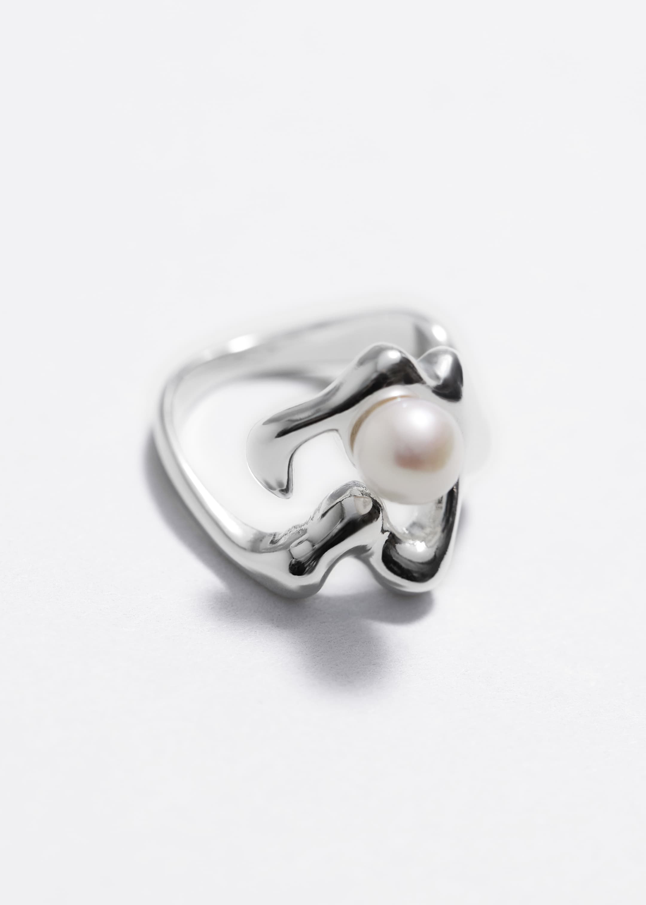 Agrandir l'image: Une bague présente une bande de métal poli de ton argenté avec un design organique et sculptural. Une perle ronde de couleur claire est nichée dans l'une des courbes fluides du métal, avec la bague inclinée pour mettre en valeur sa forme tridimensionnelle.