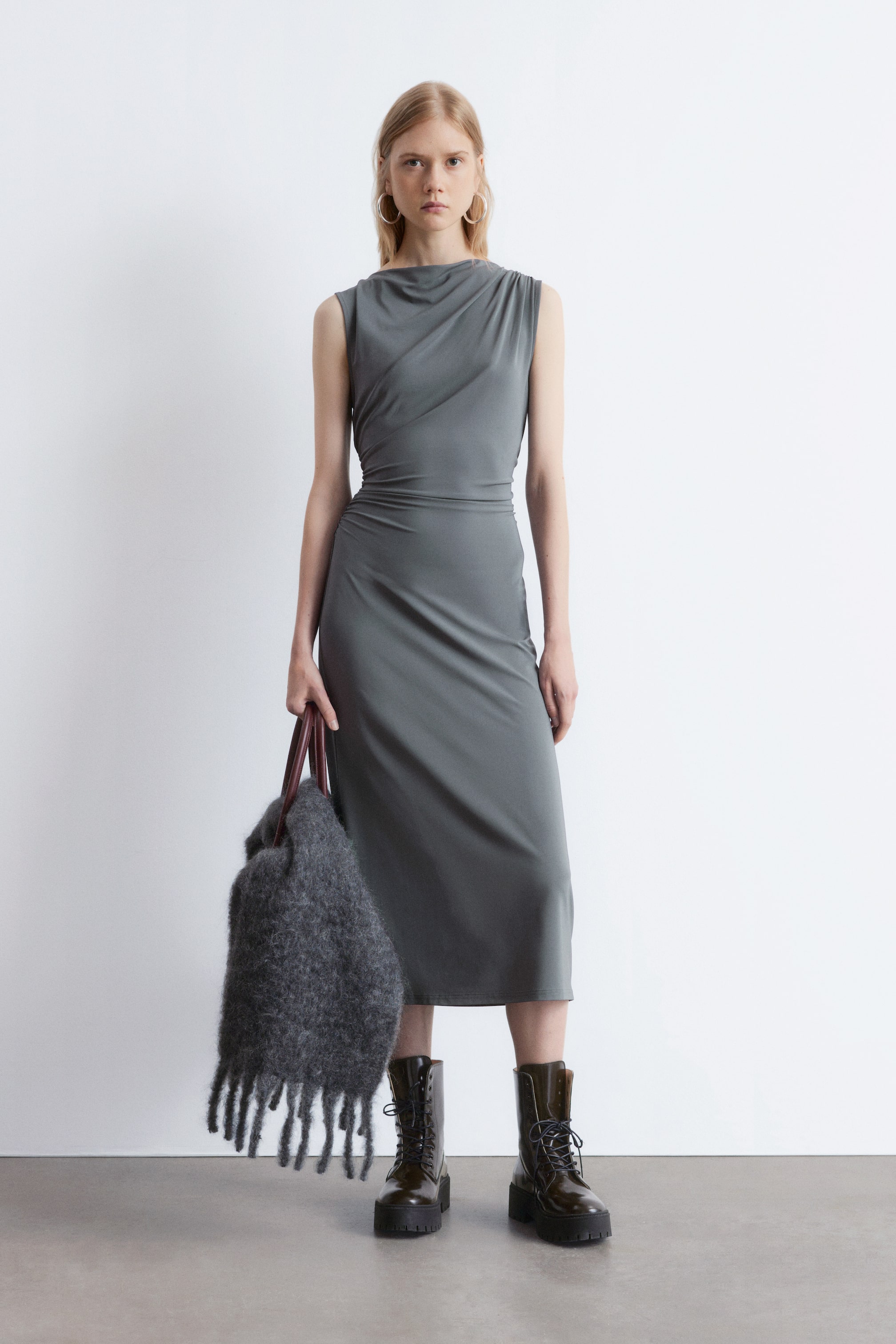 Ver imagen más grande: Un vestido midi gris sin mangas presenta un escote drapeado asimétrico y un corpiño ceñido con un sutil fruncido lateral. El vestido cuenta con una falda recta que cae hasta media pantorrilla.