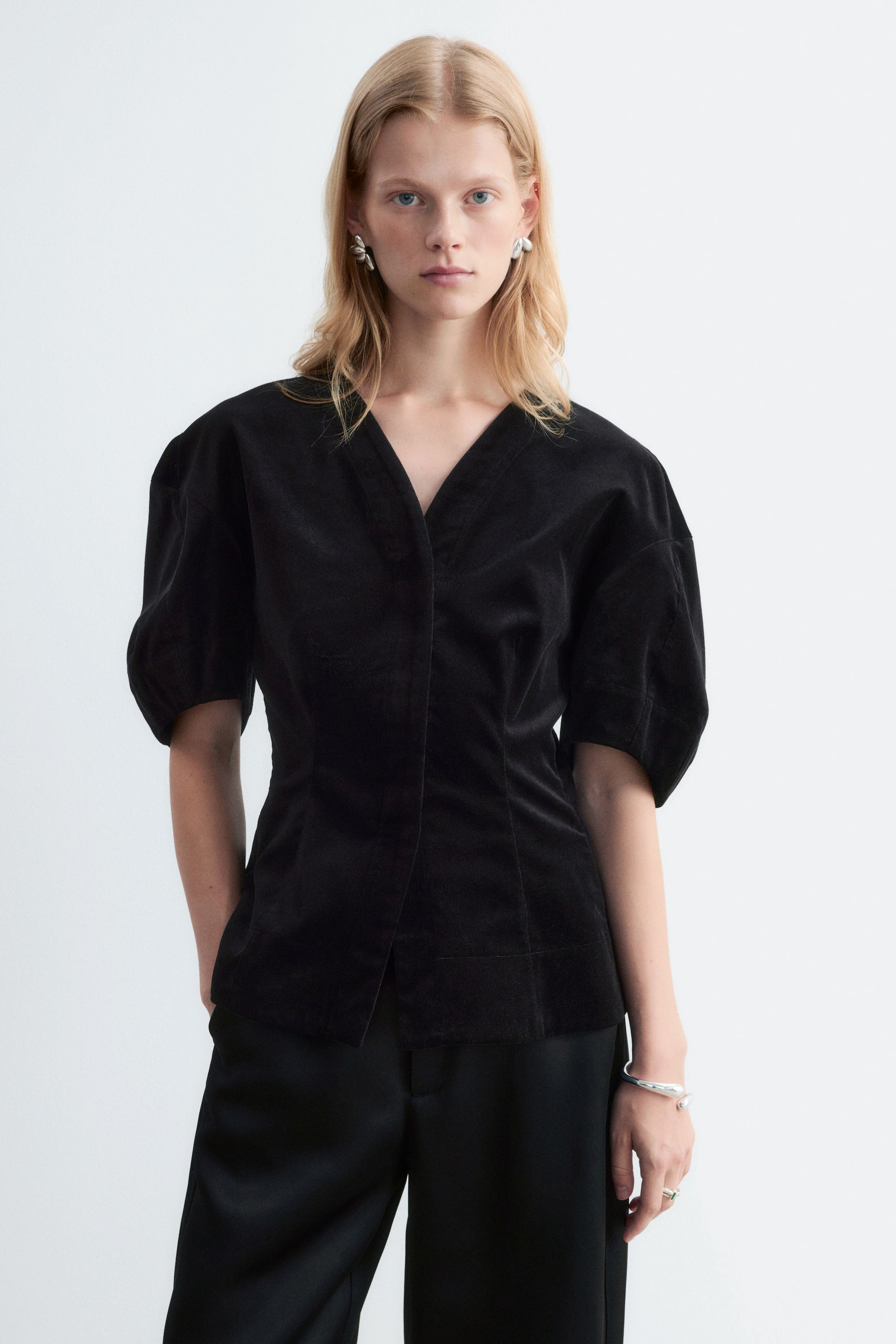 View larger image: A black velvet blouse featuring a V-neckline, a concealed front placket, and short rounded sleeves is being worn by the model. The slim-fit blouse is paired with black trousers.