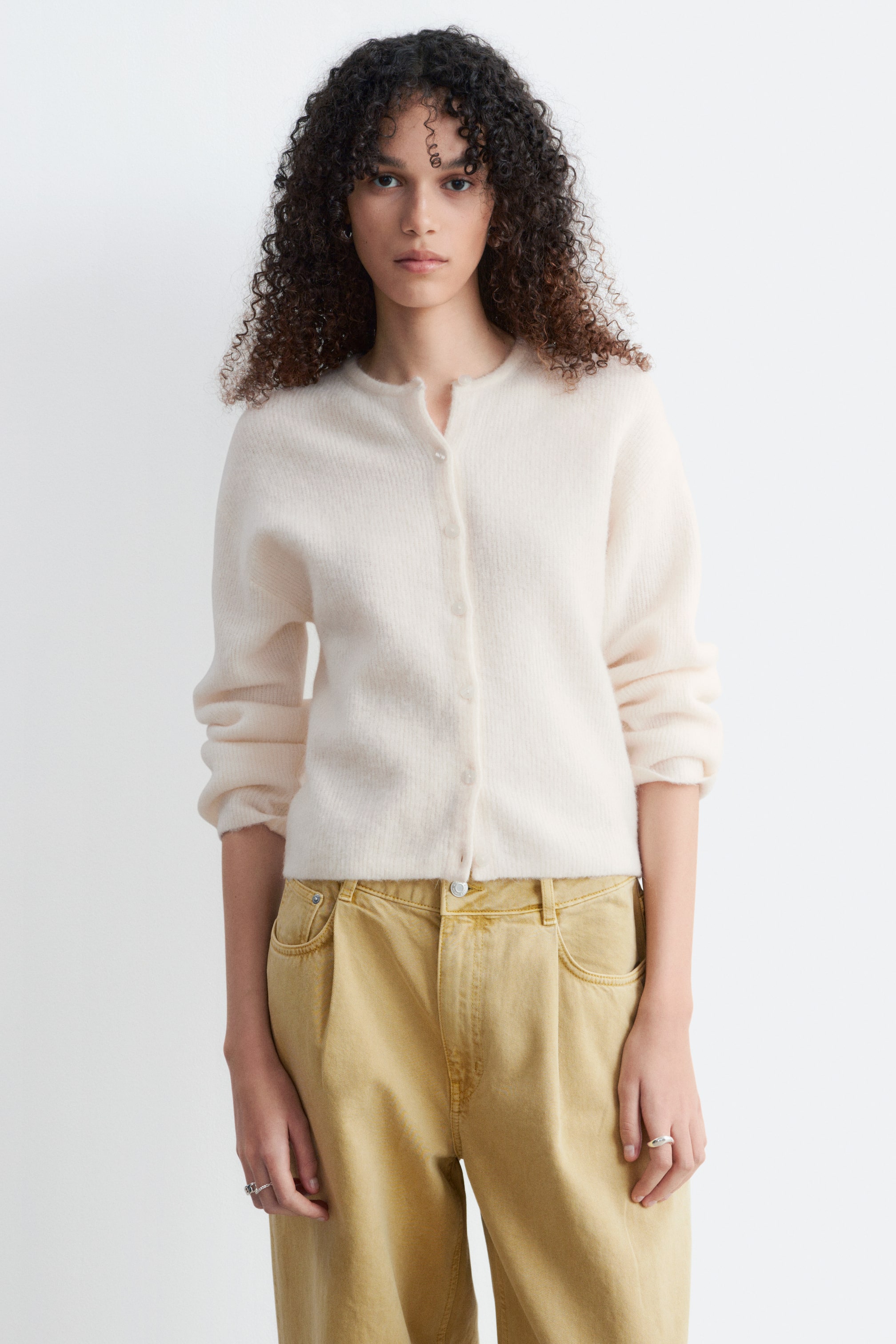 Se større billede: En off-white fuzzy strikket cardigan viser en rund halsudskæring og en frontknaplukning med lyse knapper. Den har lange ærmer med let rynkede manchetter og en lige, kort kant.