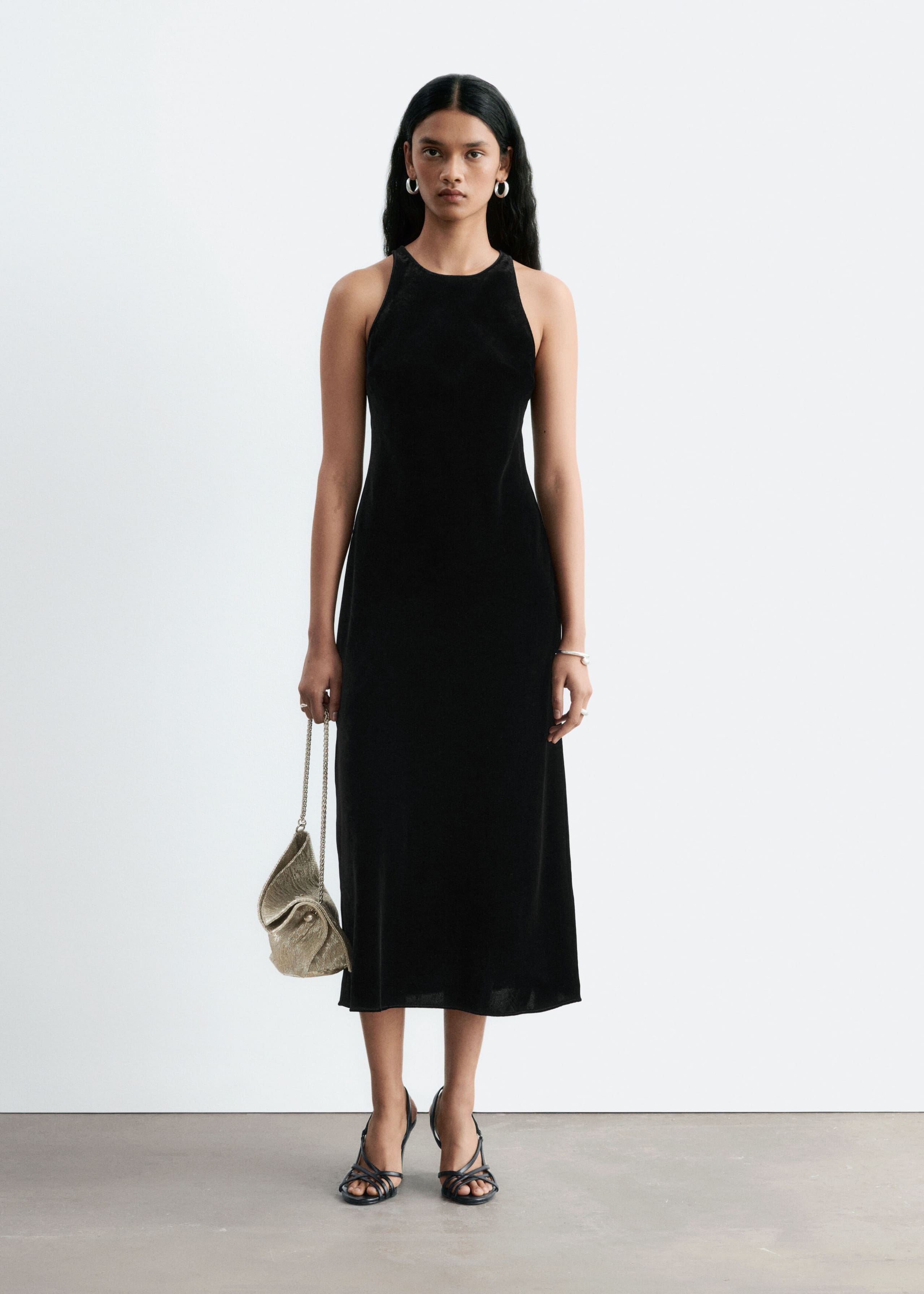 The model is wearing a black sleeveless midi dress with a round neckline, paired with a textured metallic handbag and black strappy heeled sandals.