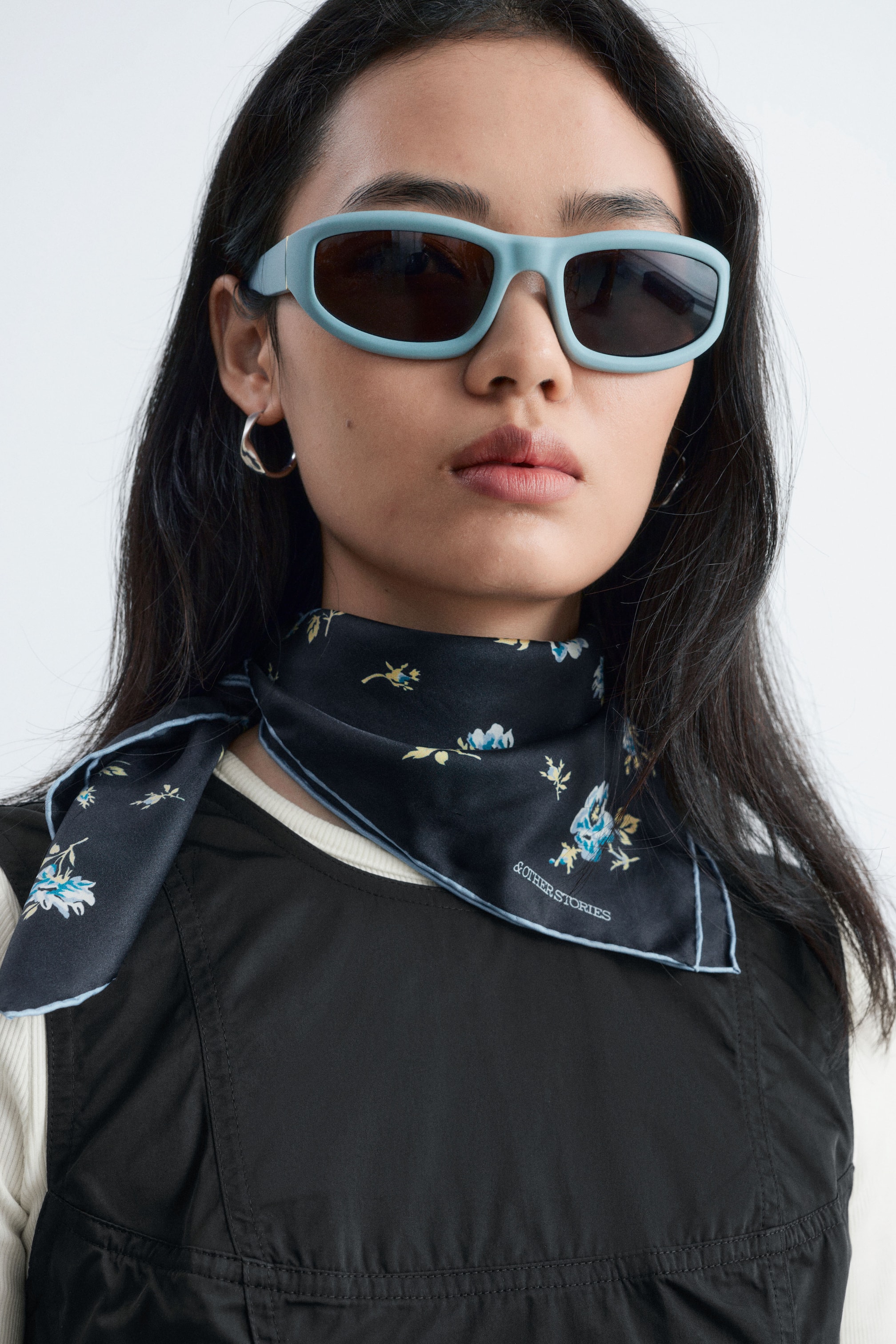 Ver imagem maior: A modelo veste um vestido mini peplum preto e um lenço de seda preto com estampado floral. O vestido sem mangas é usado sobre uma camisola canelada cor creme, enquanto o lenço quadrado está atado ao pescoço, destacando as extremidades com acabamento azul e o padrão floral.