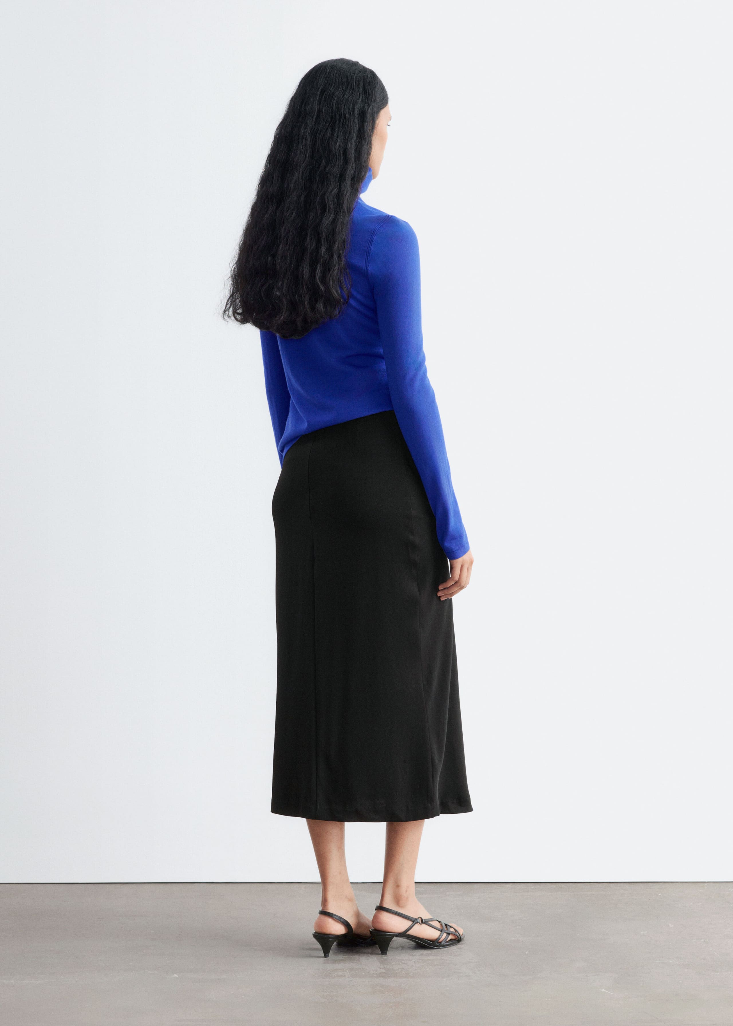 Een felblauwe merinowollen coltrui wordt gedragen door het model en heeft een slim fit, lange mouwen en een hoge hals. De top is gecombineerd met een zwarte midi-rok en zwarte sandaaltjes met bandjes en lage hak, weergegeven vanuit een driekwart achteraanzicht.