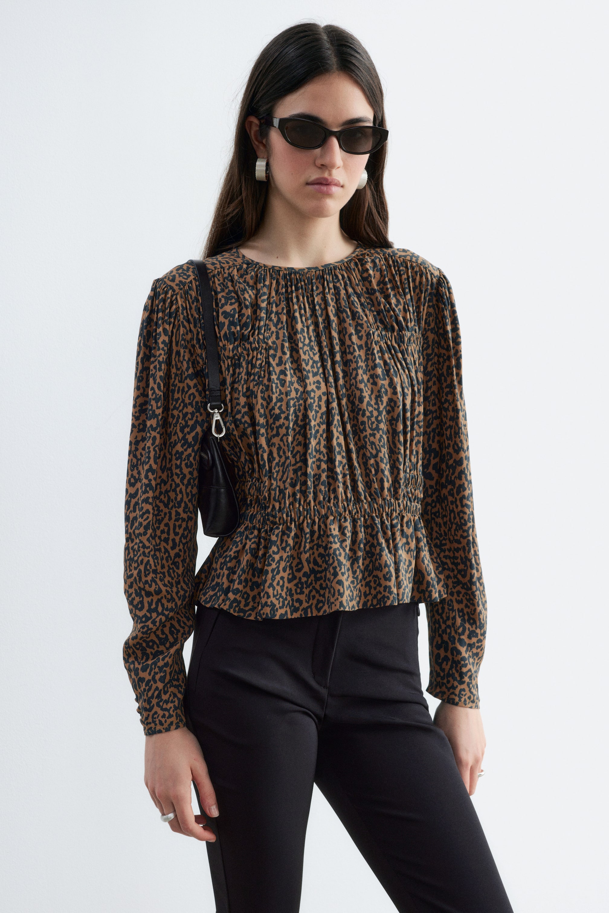 Grotere afbeelding bekijken: Een blouse met luipaardprint, voorzien van gerimpelde smockdetails over de borst en een elastische taille, wordt gedragen door het model. De top met lange mouwen is gestyled met een zwarte broek en een zwarte schoudertas.