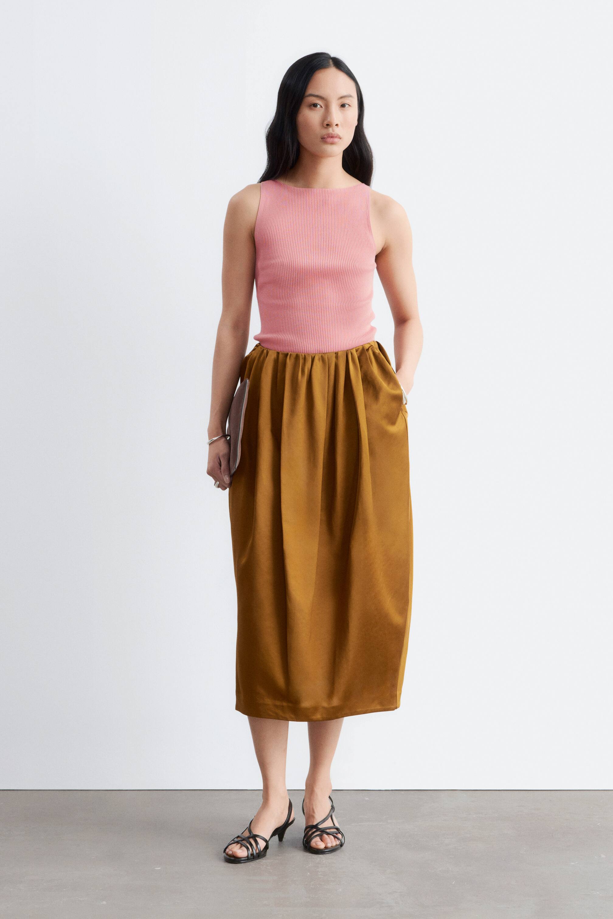 Visa större bild: En figurnära rosa ribbstickad linnepresenteras, med en båtringning. Den bärs instoppad i en voluminös gyllenbrun midikjol, som visar upp en rynkad elastisk midja och sidofickor.