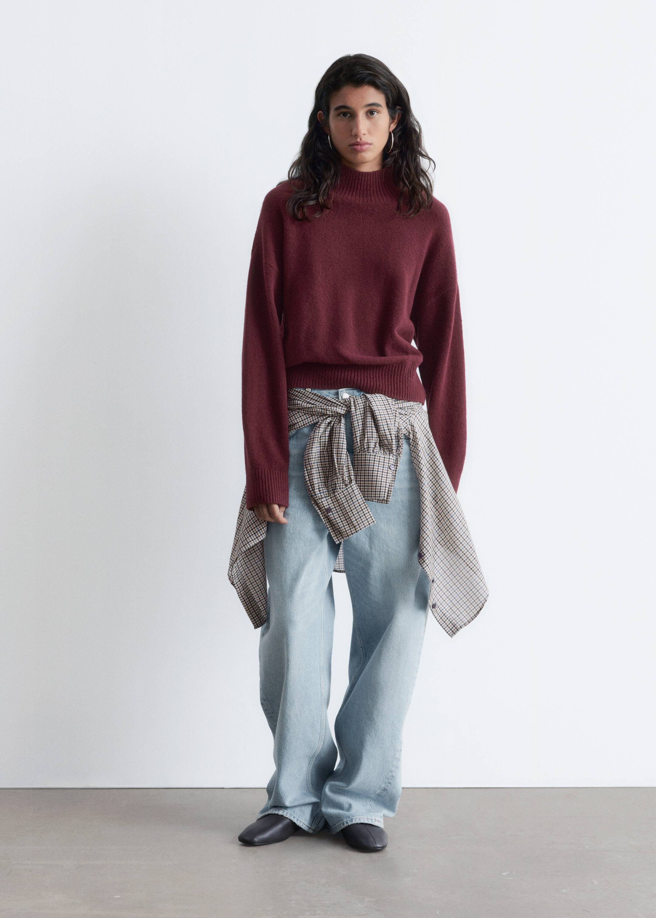 View larger image: A burgundy mock-neck sweater with ribbed cuffs and hem is being worn with light wash wide-leg jeans. A brown and cream plaid shirt is being tied around the waist, and dark flat shoes are completing the outfit, which is presented from a front-facing angle.