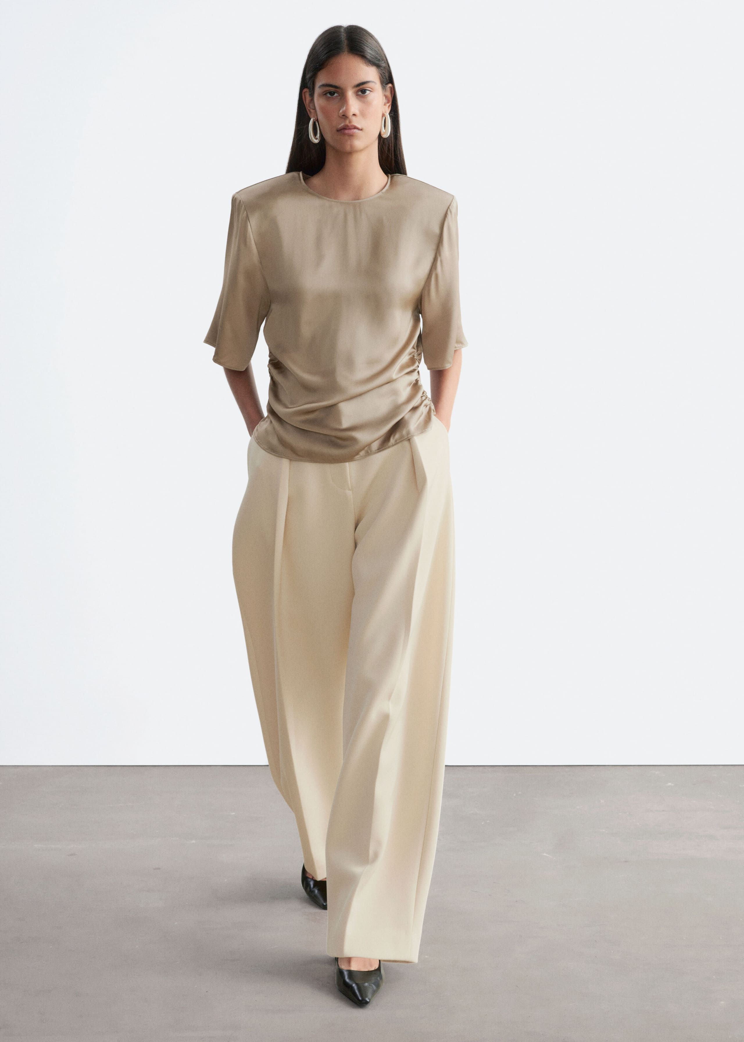 A beige satin-finish blouse with structured shoulders, short sleeves, and gathered side pleating is worn by the model. The blouse is paired with cream-colored wide-leg trousers.