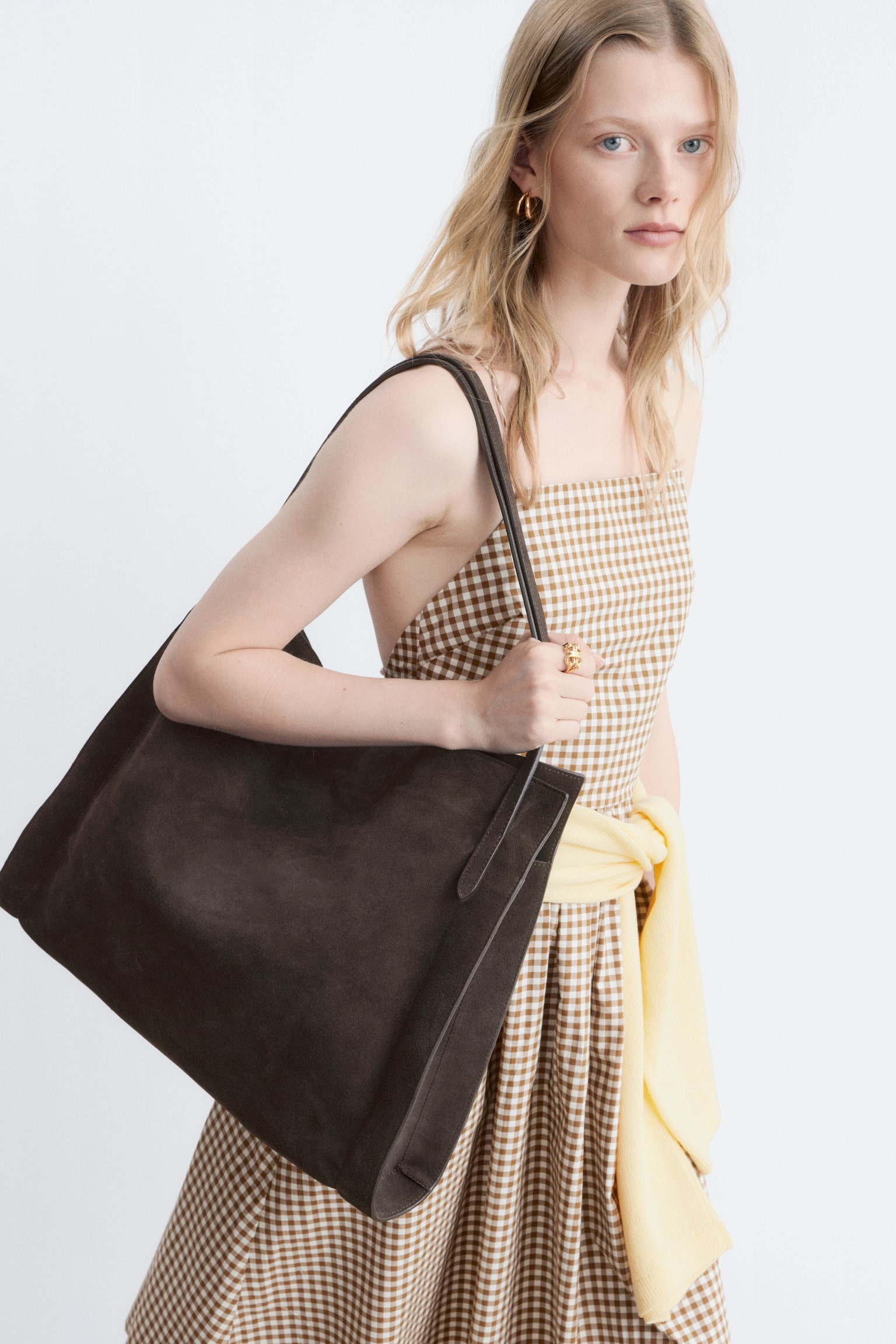 A dark brown suede tote bag is hanging from the shoulder and being held by one hand, displaying its rectangular shape and two slender straps. - 1