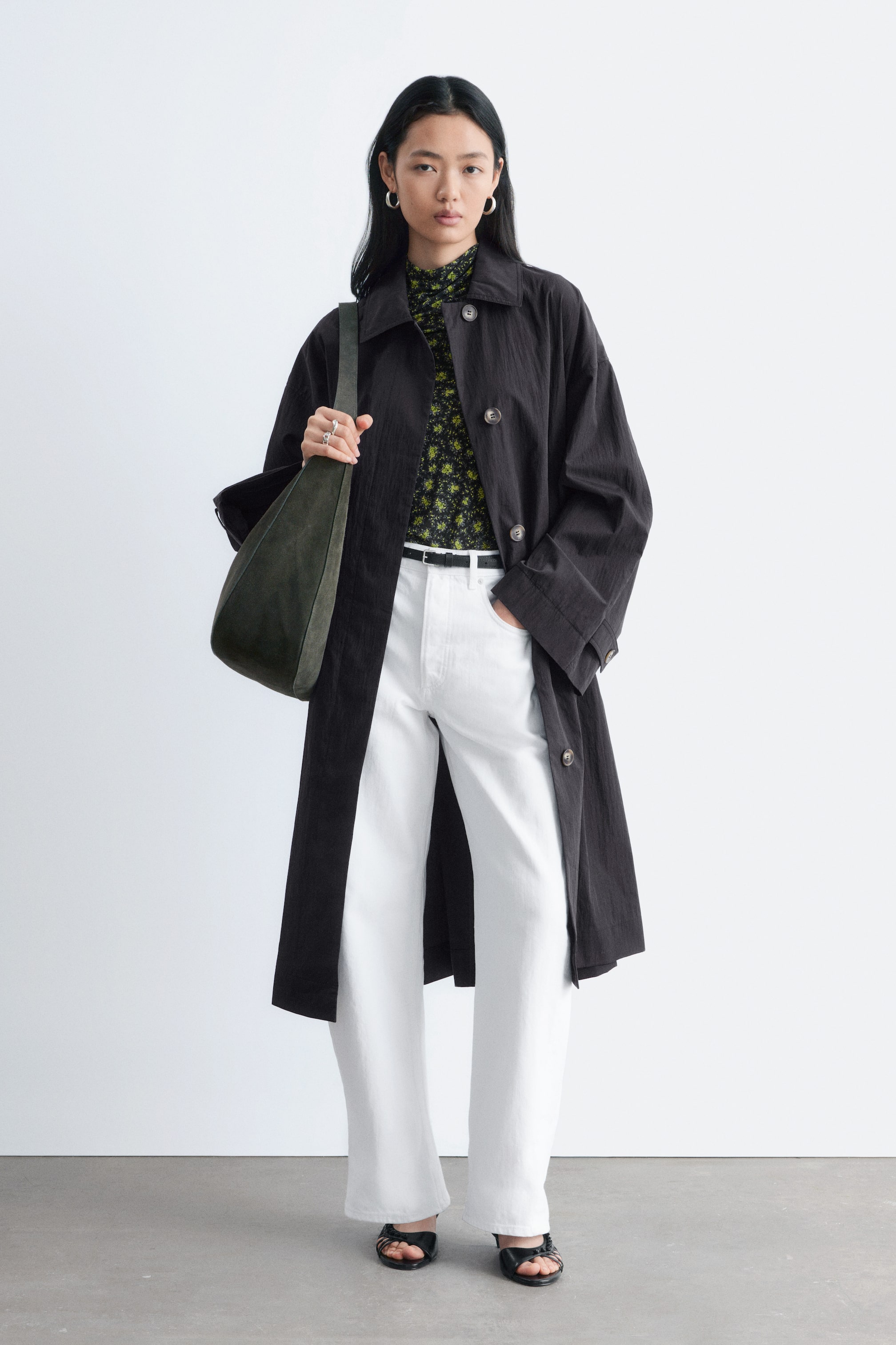 Visualizza immagine più grande: Un cappotto nero al ginocchio con colletto classico e silhouette ampia è indossato dalla modella sopra una camicia a fiori e pantaloni bianchi. Bottoni scuri sono visibili lungo l'apertura frontale del cappotto, e la modella è in piedi con una mano in una tasca laterale a filetto.