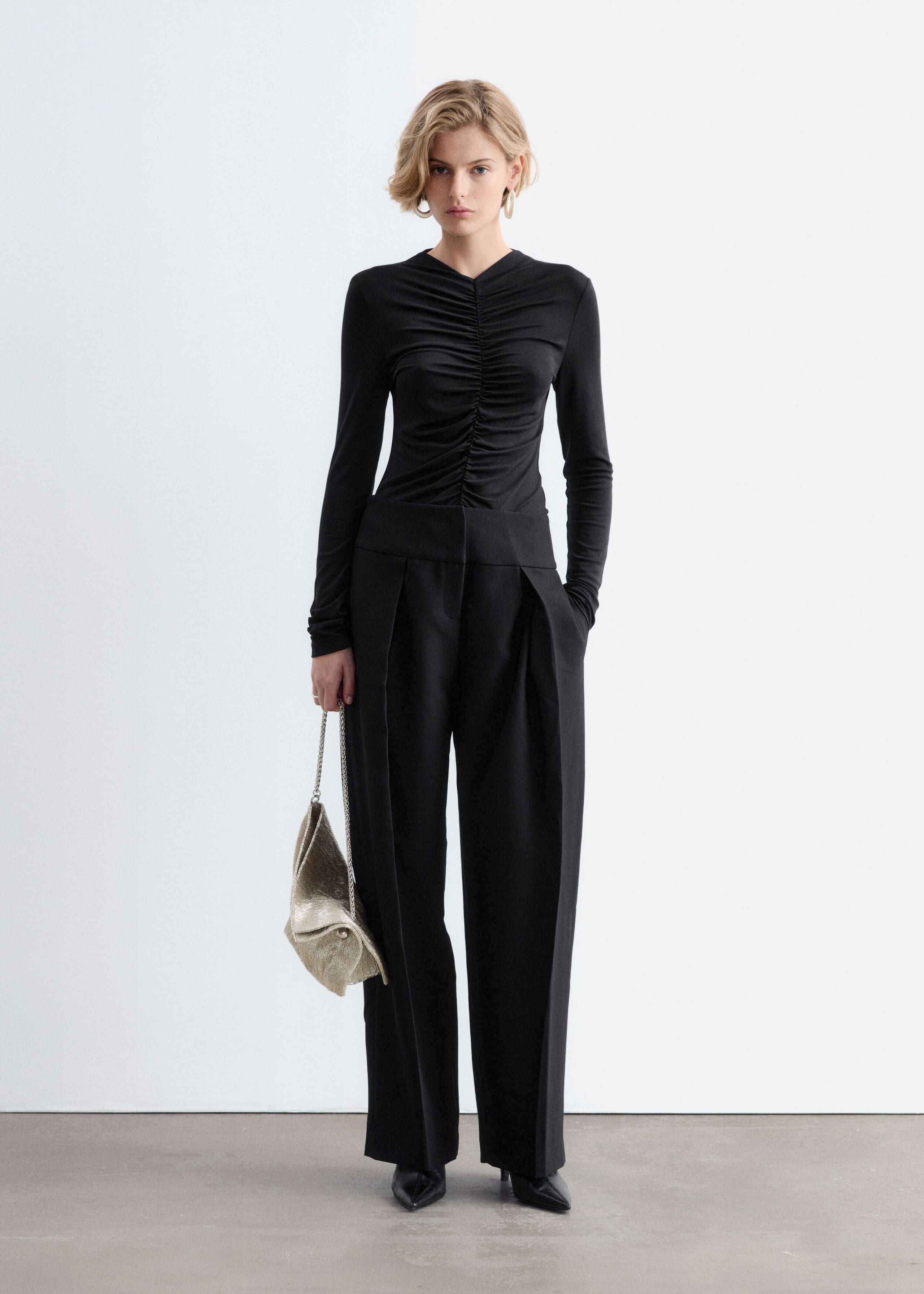 A black long-sleeve top with prominent gathered detailing along the center-front seam is worn by the model. The slim-fitting top is styled tucked into wide-leg black pants.