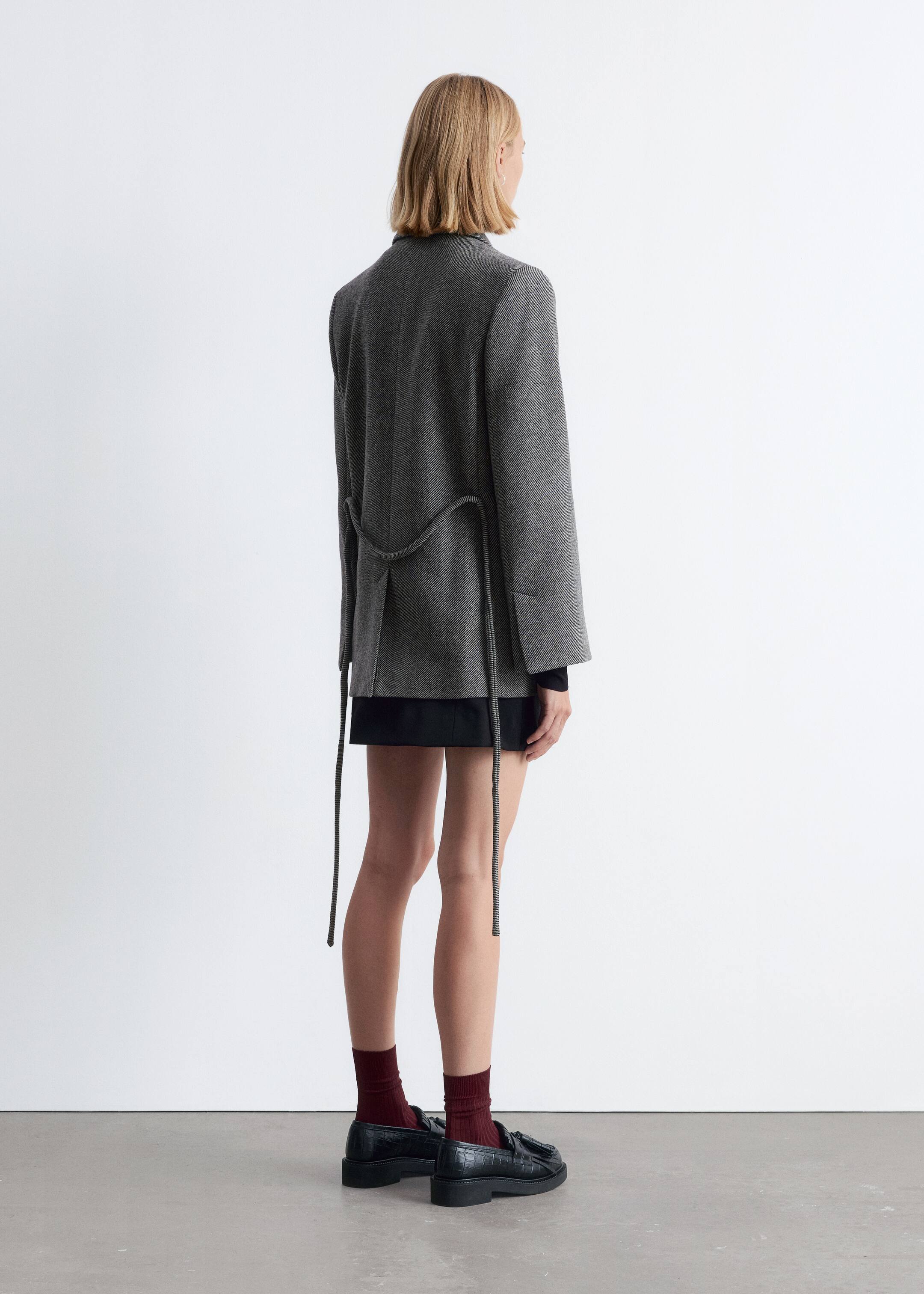 Agrandir l'image: Un blazer gris est vu de dos, porté sur un vêtement noir, mettant en valeur sa coupe ample et décontractée. Il présente un motif subtil en chevrons, et un fin cordon tressé est drapé lâchement à l'arrière, avec de longues extrémités pendantes.