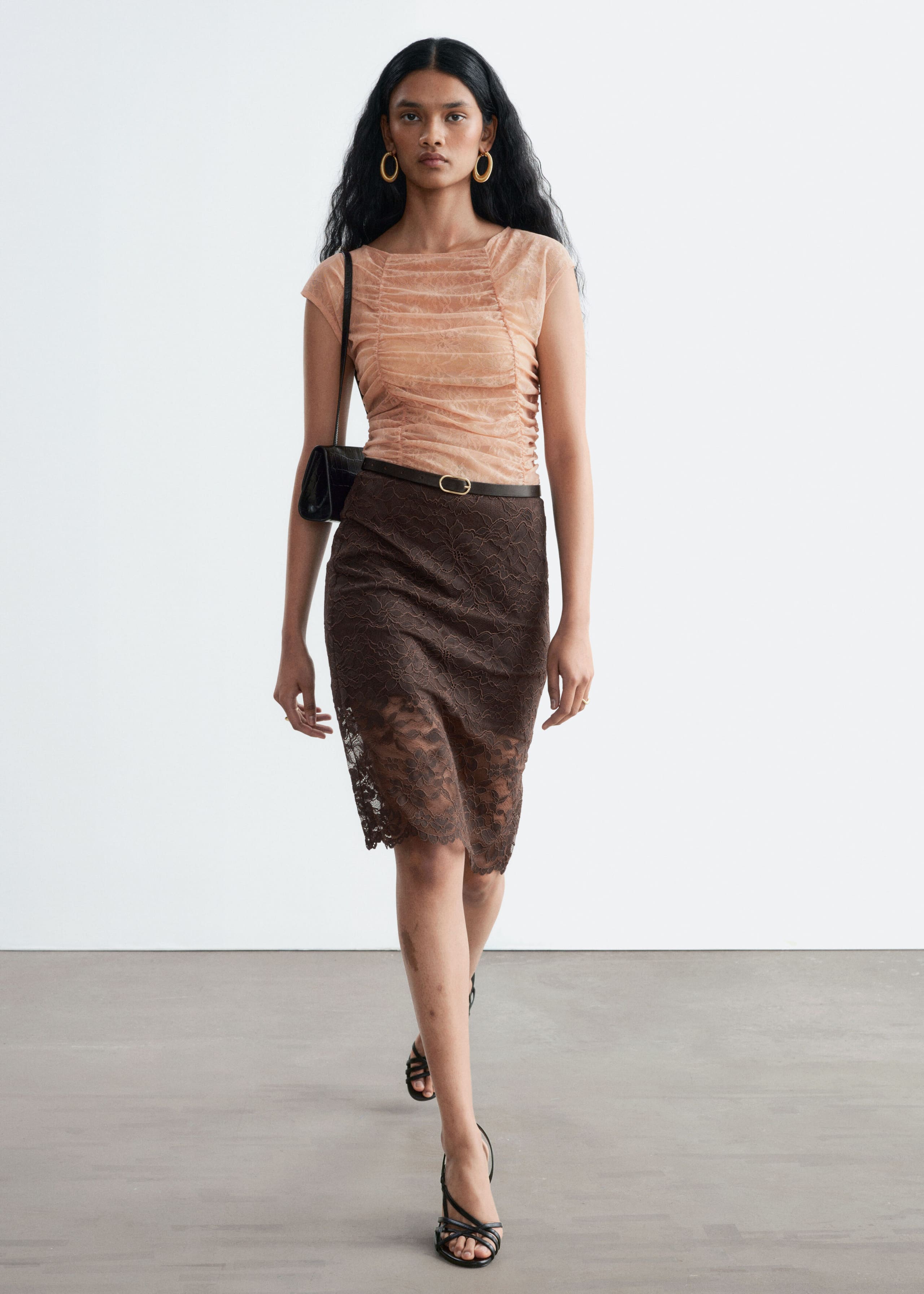 A peach-coloured lace top featuring a boat neckline, cap sleeves, and ruched detailing is being worn by the model. The top is tucked into a dark brown lace skirt styled with a thin belt.