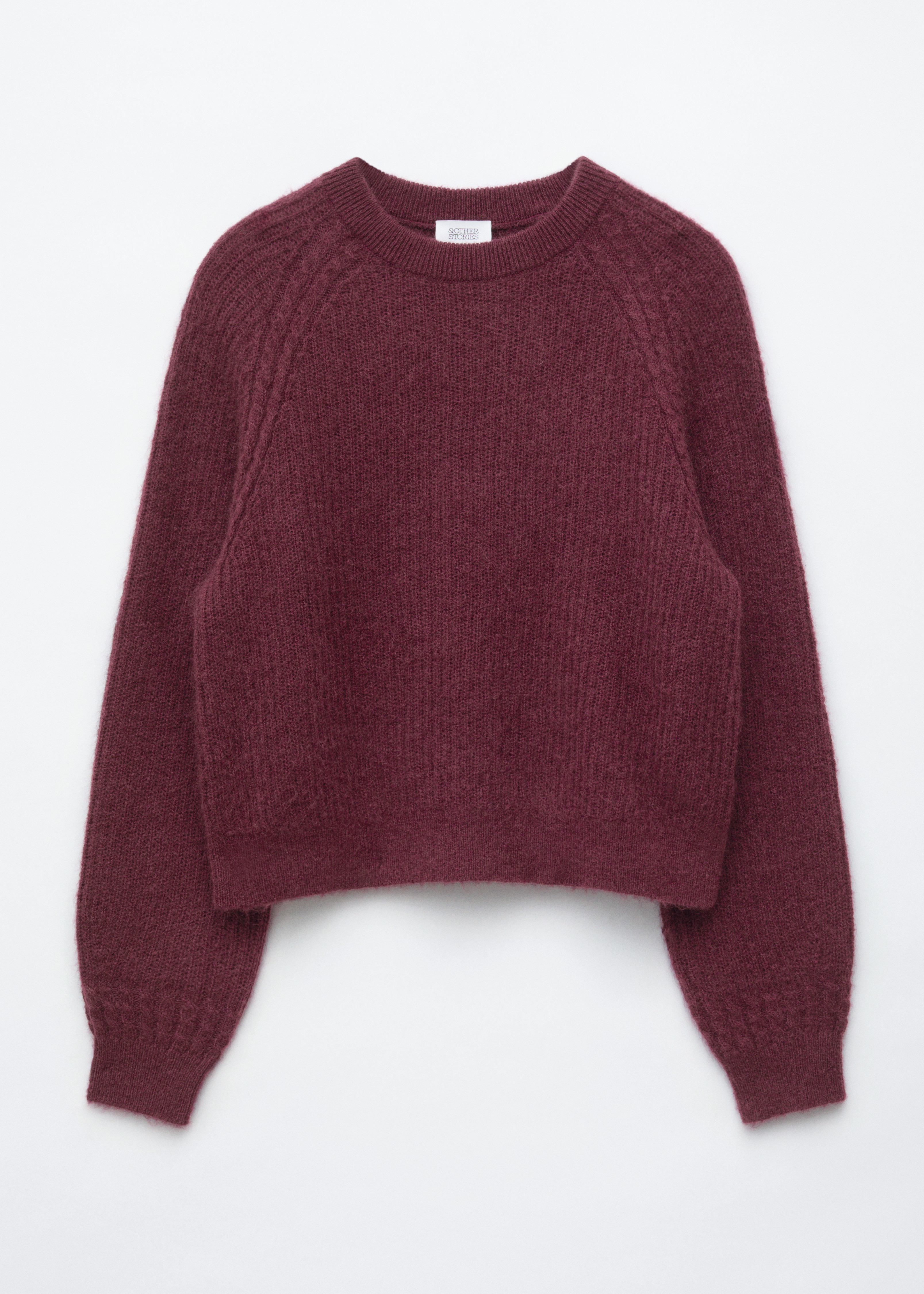 Een bordeauxrode trui van mohairmix met geribde textuur, ronde hals en raglanmouwen wordt gedragen door het model. De losvallende trui is gestyled met een zwarte jeans en een donkere schoudertas.