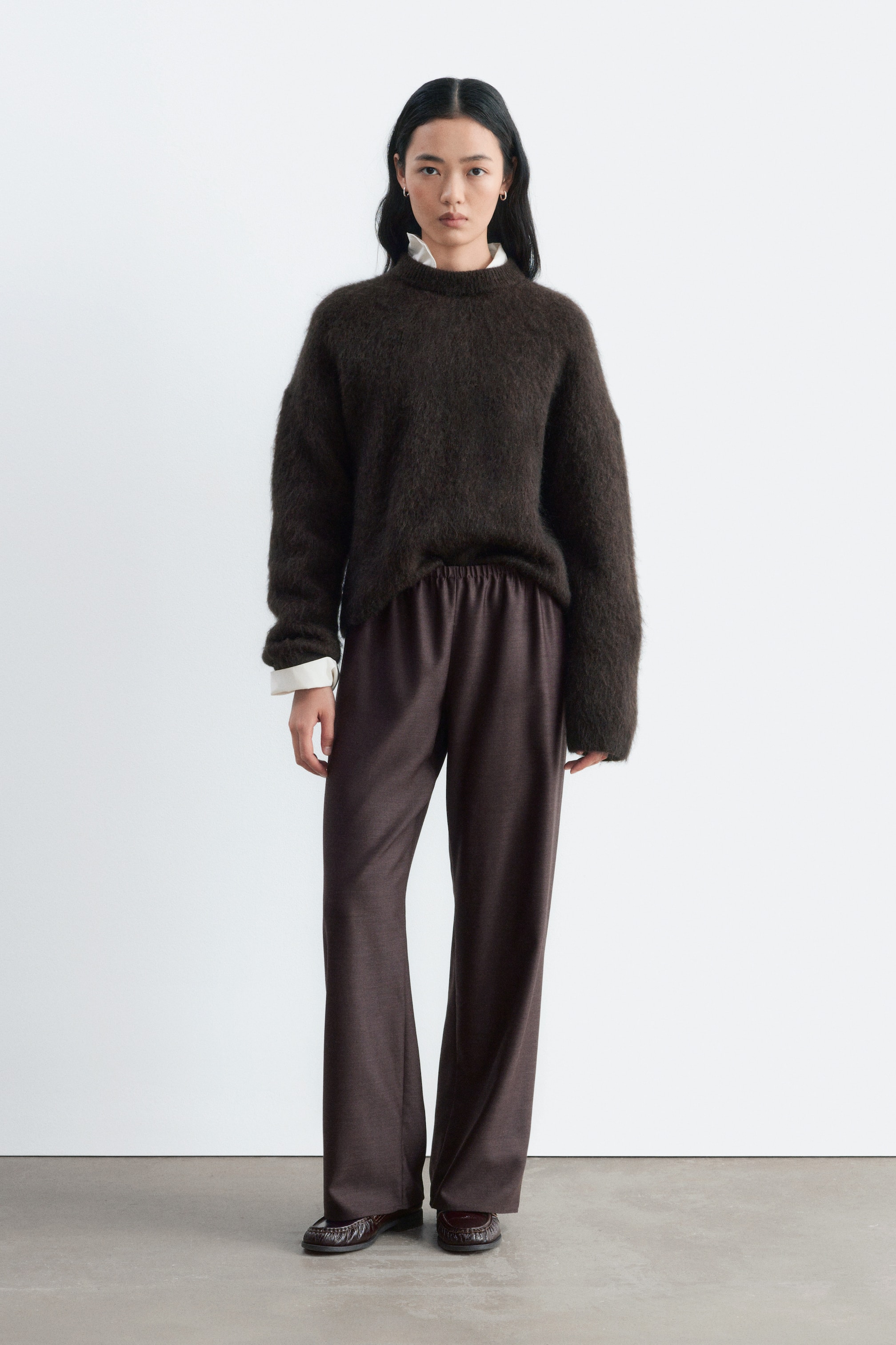 Visualizza immagine più grande: Pantaloni di lana marrone scuro con vita elasticizzata e silhouette a gamba dritta, indossati dalla modella. I pantaloni sono abbinati a un maglione testurizzato marrone scuro, sovrapposto a una camicia bianca con colletto.