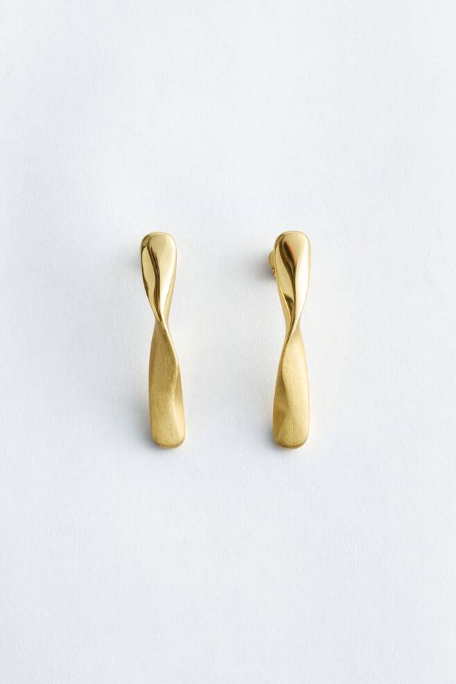 A pair of gold-toned earrings featuring an elongated, twisted design. The top section of each earring appears polished, transitioning into a brushed finish along its twisted, wider lower part, as they lie flat and face forward. - 1