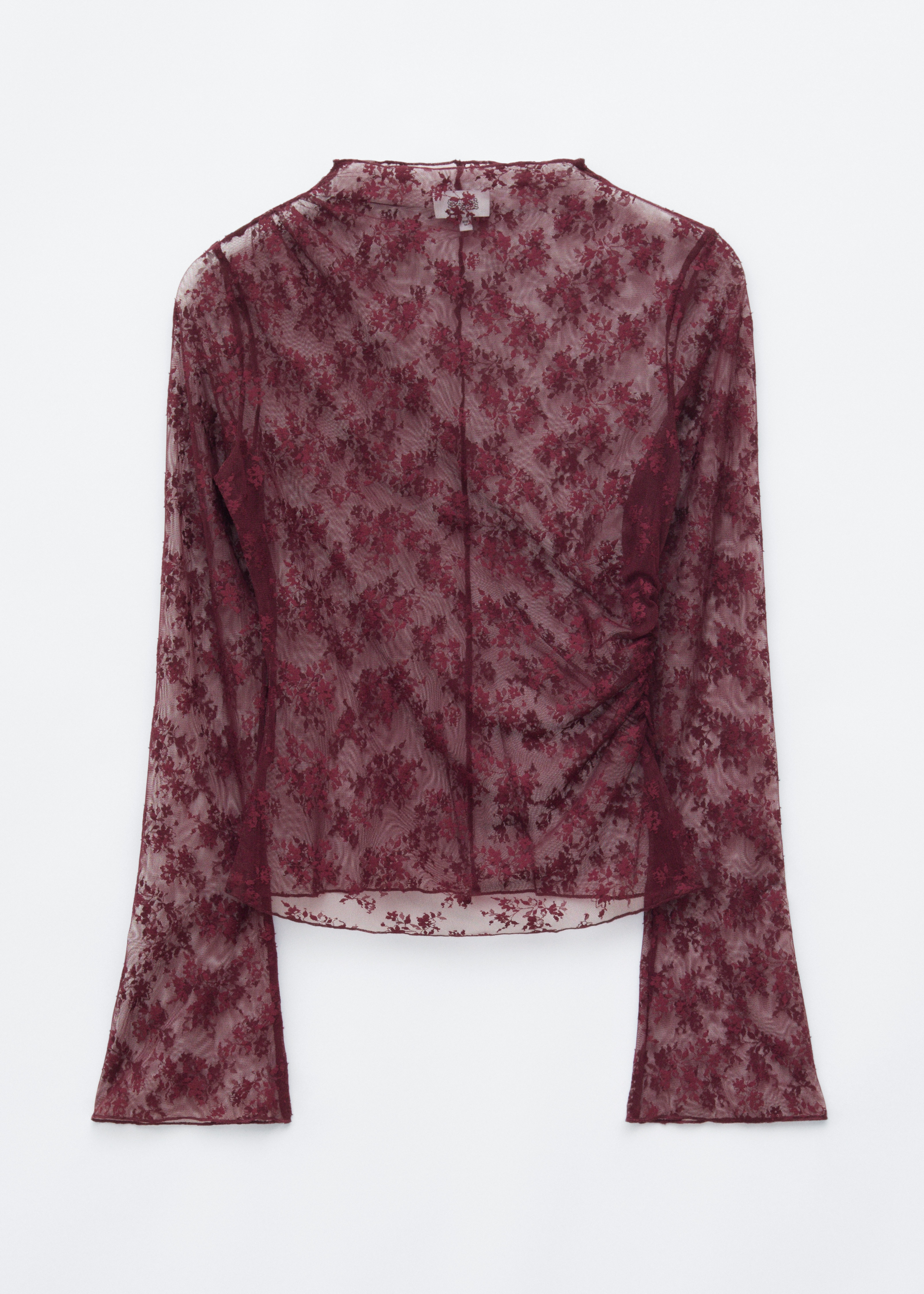 Un haut en dentelle florale bordeaux transparente Ã  col montant et manches longues Ã©vasÃ©es est portÃ© par le mannequin par-dessus un soutien-gorge assorti. Le haut est rentrÃ© dans une jupe en cuir bordeaux, et le mannequin tient une grande pochette gris foncÃ©.
