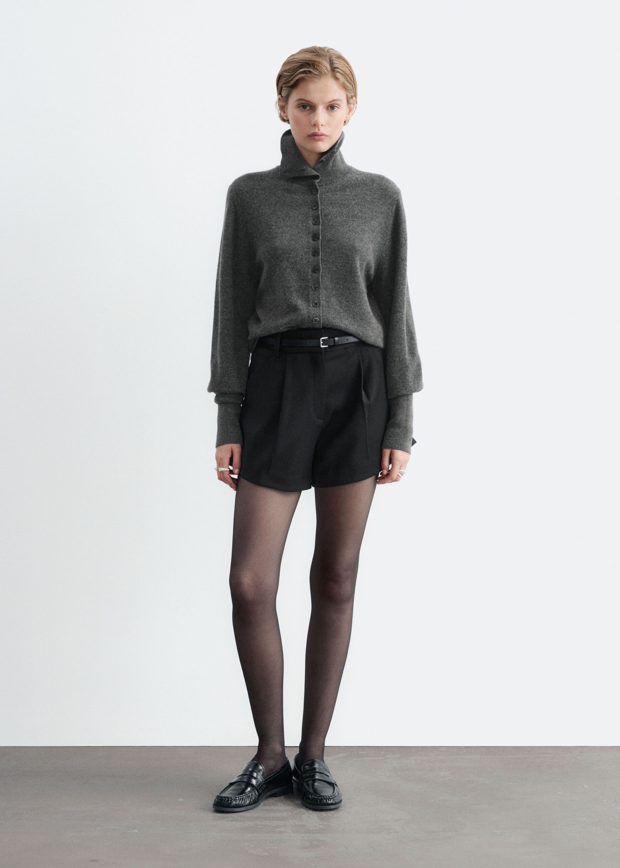 Black high-waisted shorts with front pleats and an A-line silhouette are worn by the model, secured with a slim black belt. The shorts are styled with a tucked-in grey high-neck knit jumper, sheer black tights, and black leather loafers.