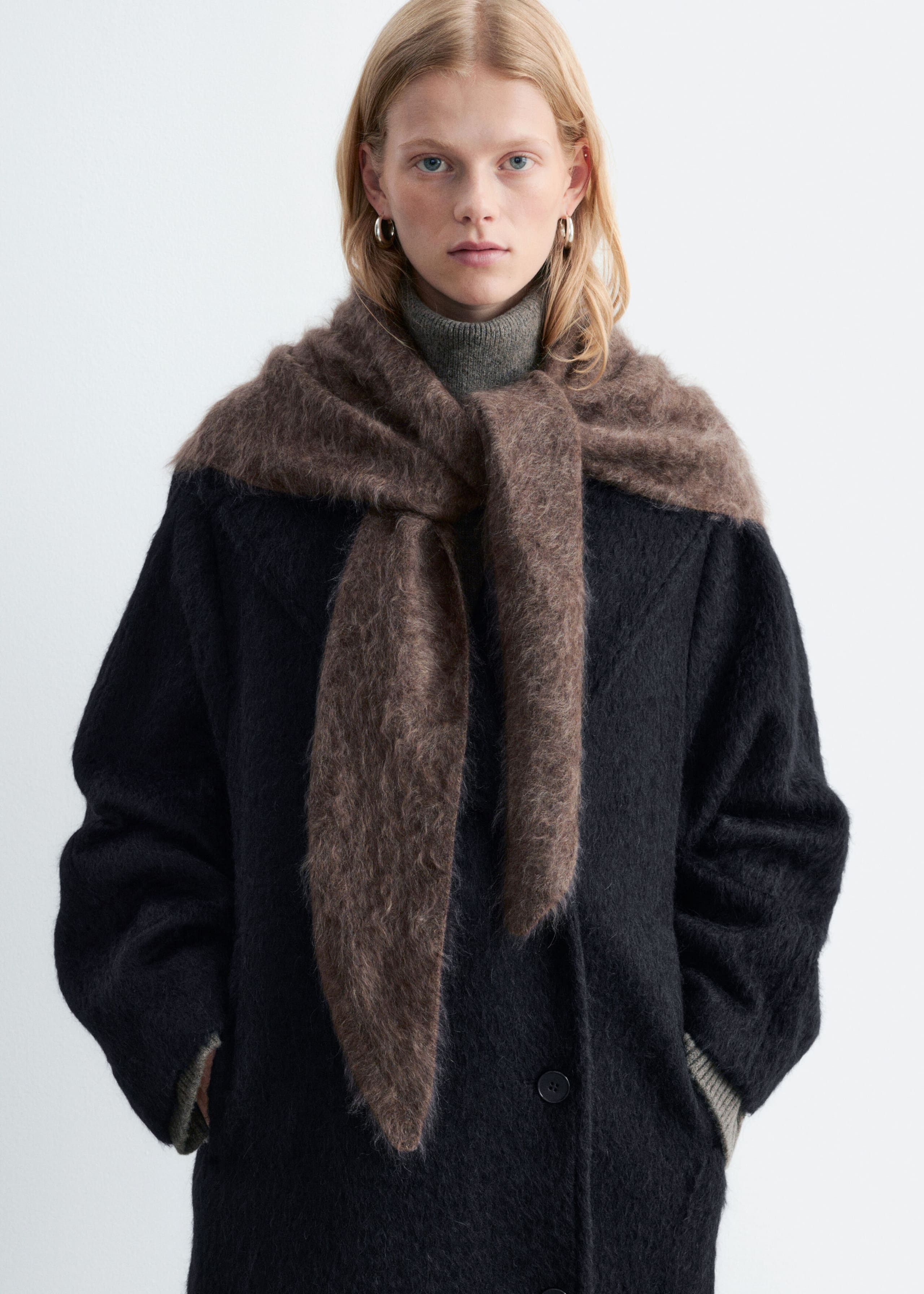 A brown brushed cashmere triangle scarf is worn by the model, draped over the shoulders with the ends loosely tied at the front. The textured scarf is layered over a black coat and a gray turtleneck sweater.