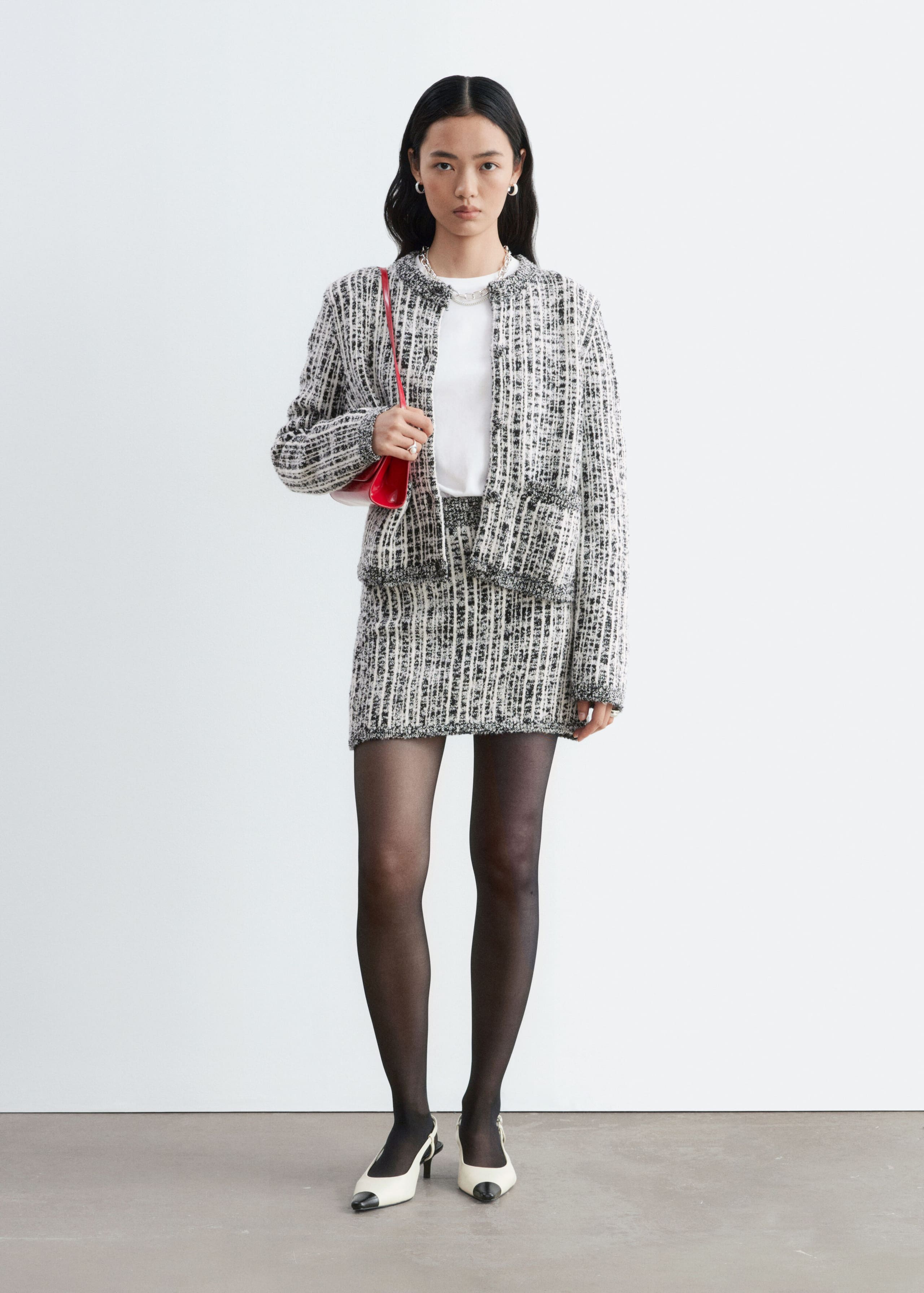 The model is wearing a black-and-white textured tweed jacket with a matching mini skirt. The open-front jacket is layered over a white T-shirt, and a red shoulder bag is tucked under the arm.