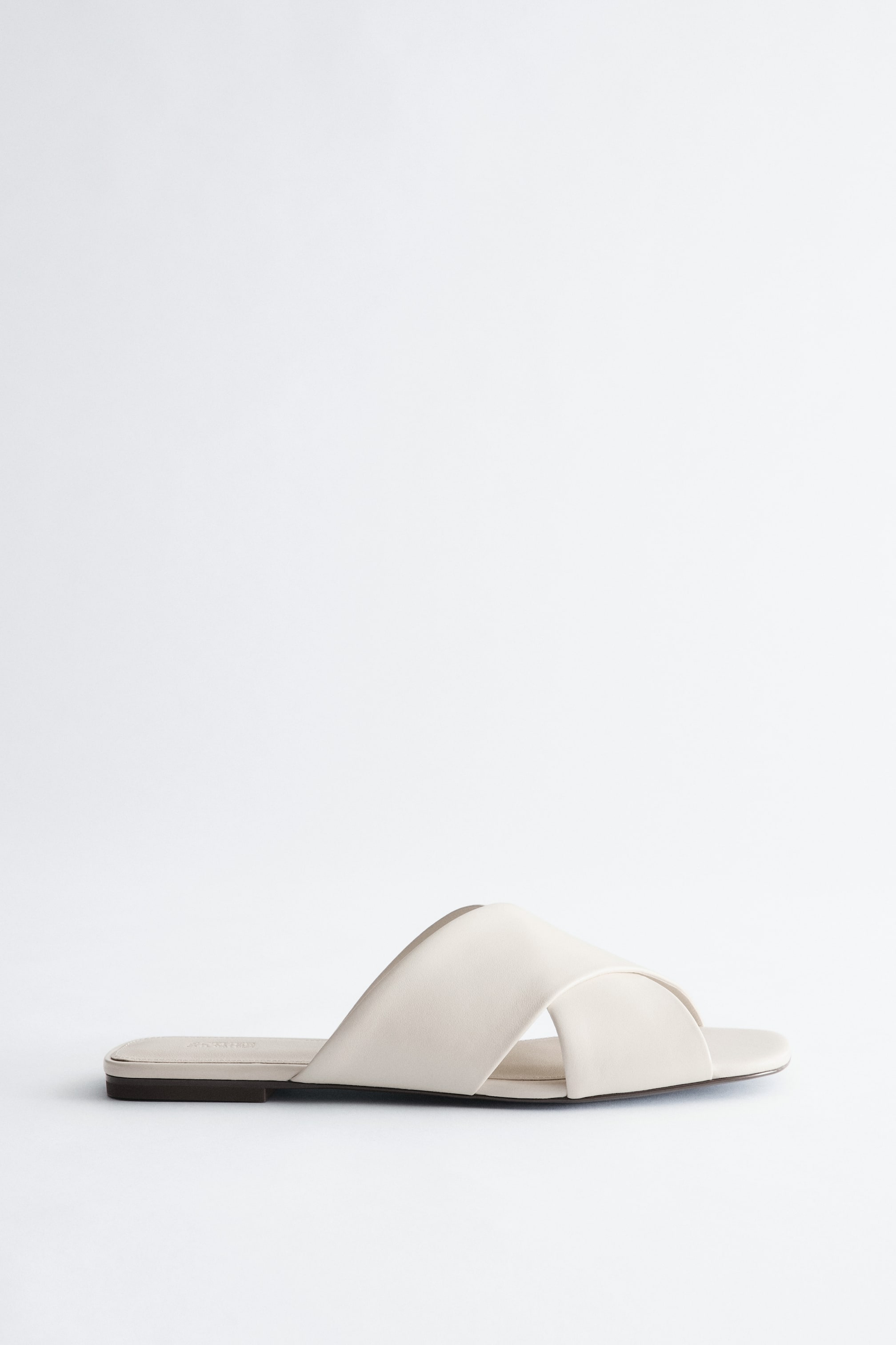 Agrandir l'image: Une sandale à enfiler beige clair est orientée vers la gauche, mettant en valeur une semelle plate et deux larges brides volumineuses formant un design croisé sur le dessus.