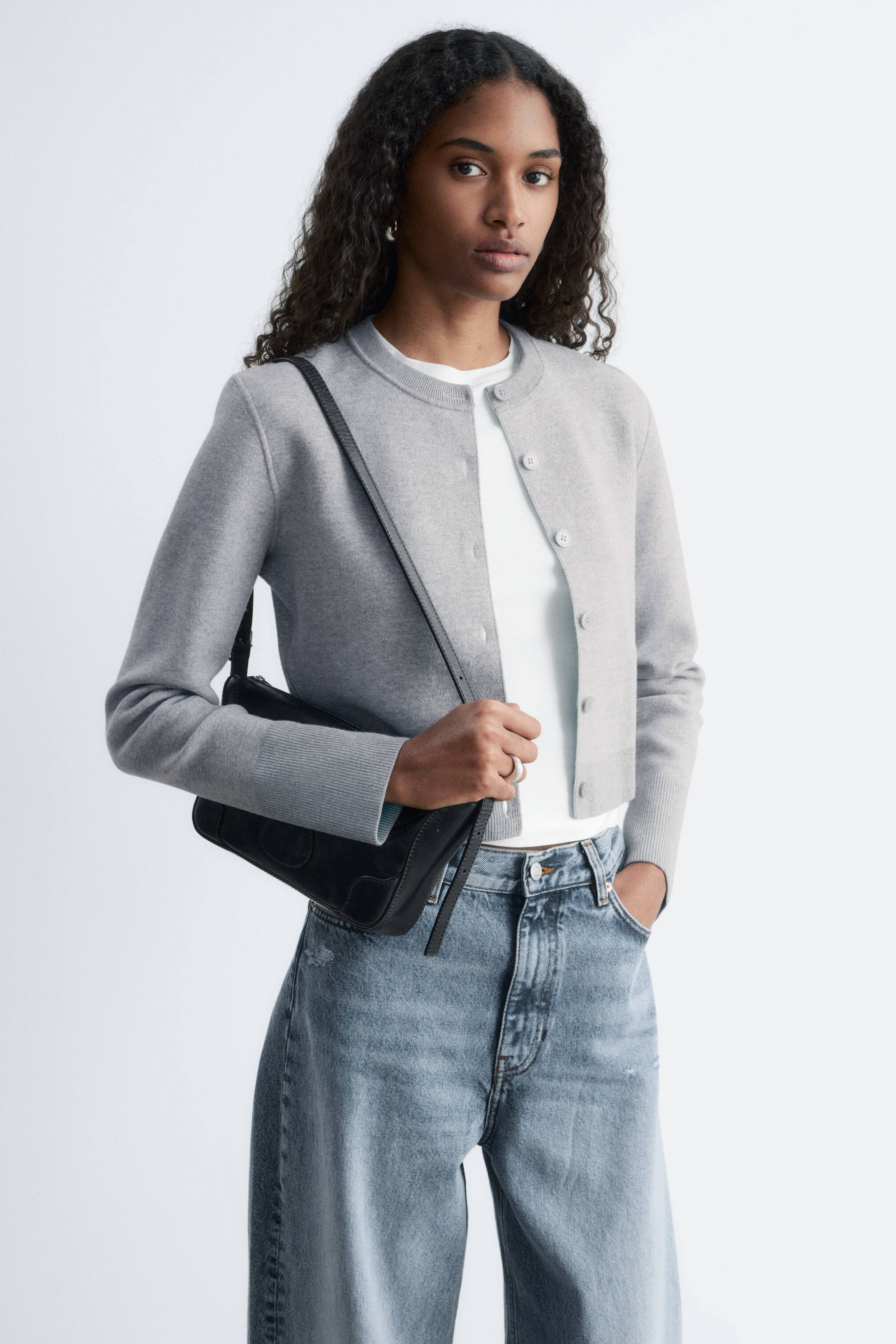 Agrandir l'image: Un cardigan gris clair est porté, avec un col rond et une fermeture à boutons sur le devant avec des boutons de couleur claire. Il présente des manches longues avec des poignets côtelés et un ourlet côtelé, et est superposé sur un t-shirt blanc.