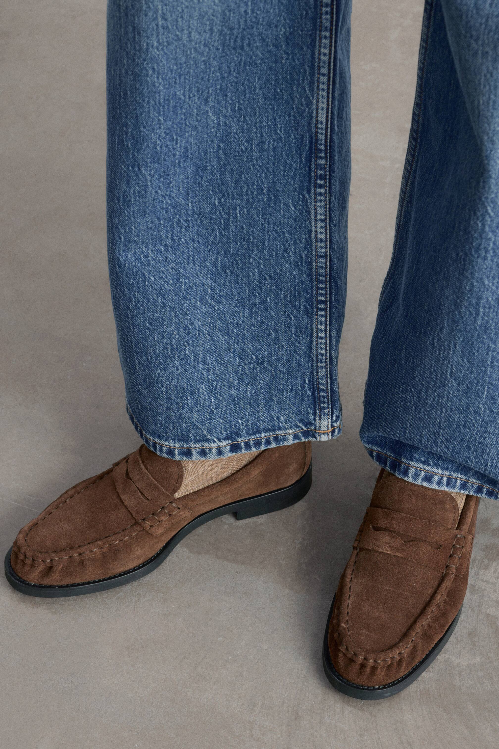 Größeres Bild anzeigen: Braune Penny-Loafer aus Spaltsuede werden getragen, wobei der linke Schuh nach links zeigt, um sein Profil und den flachen Absatz zu zeigen. Die Loafer sind mit einem klassischen Sattelriemen über dem Vorderblatt und sauberer Schürsennaht gestaltet und werden zu weit geschnittenen blauen Jeans kombiniert.