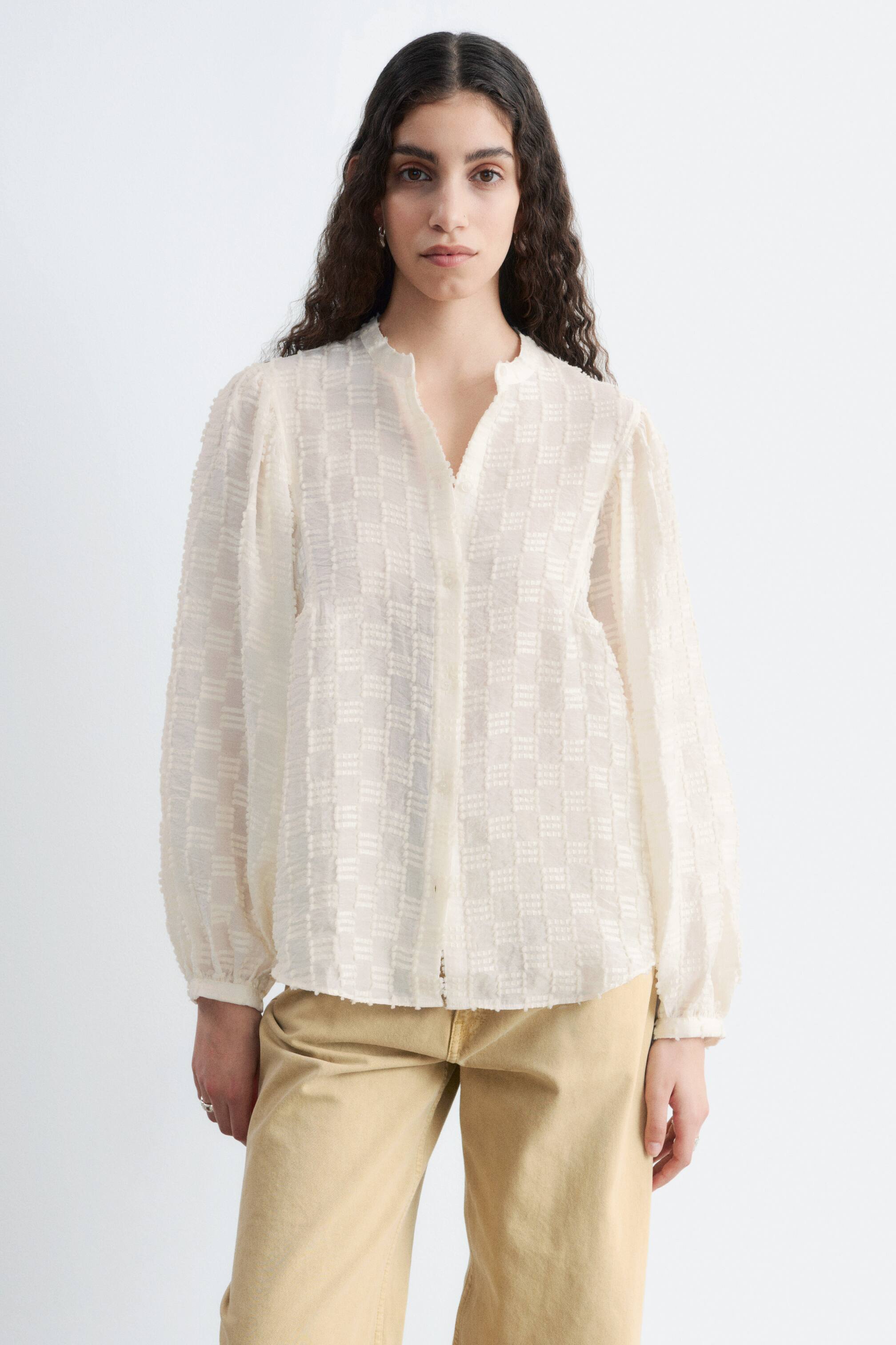 Grösseres Bild ansehen: Eine cremefarbene Bluse wird von vorne präsentiert, mit einem strukturierten Karomuster. Sie ist mit einem Stehkragen, einem V-Ausschnitt mit teilweise verdeckter Knopfleiste und langen Puffärmeln gestaltet, die in ein-Knopf-Manschetten zusammenlaufen.