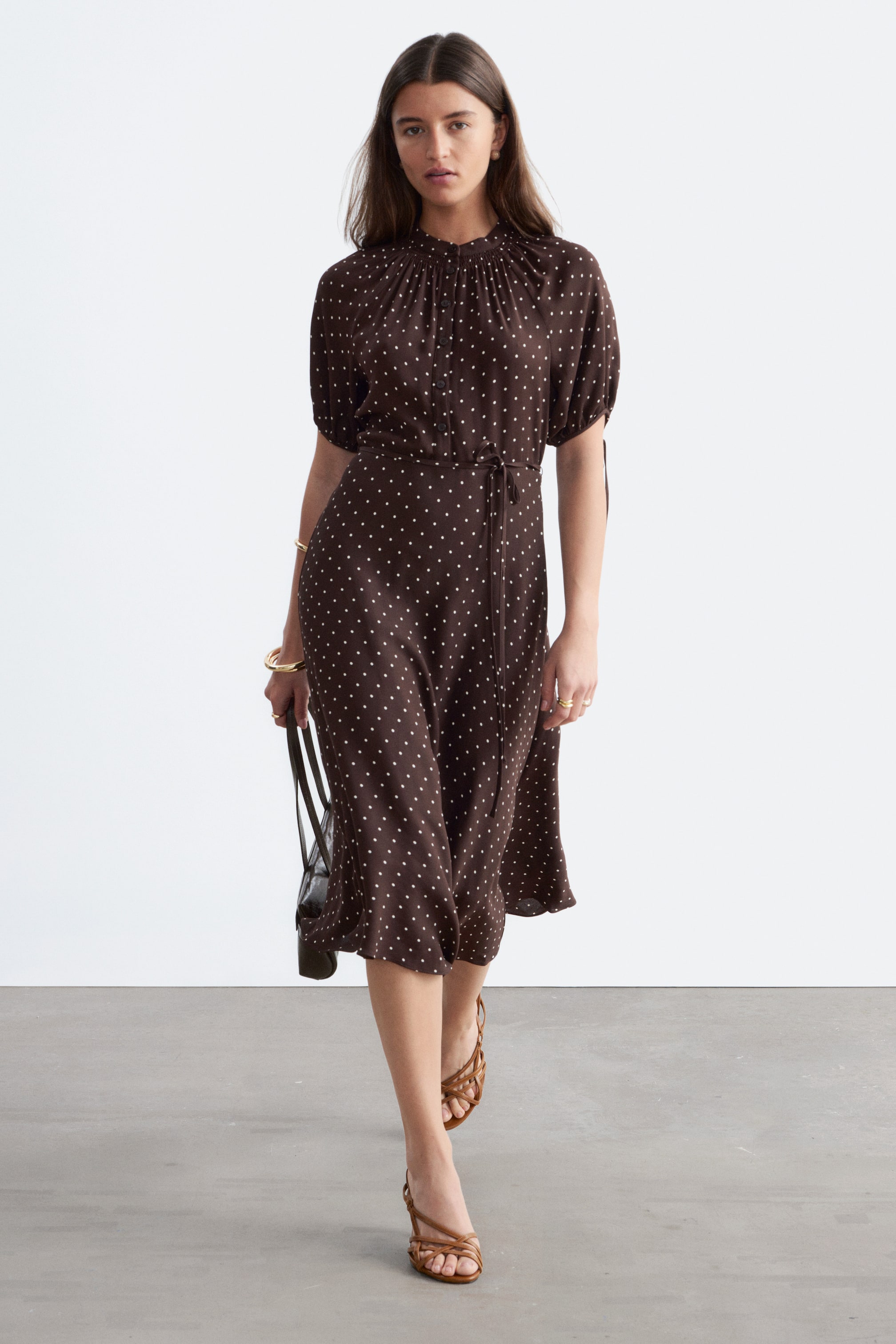 Se større billede: En brun midikjole i blød satin med hvide polkaprikker bæres af modellen. Den er designet med en rynket høj halsudskæring, korte pufærmer med bindemanchetter og et bindebånd i samme stof, der fremhæver taljen og skaber en flydende silhuet.