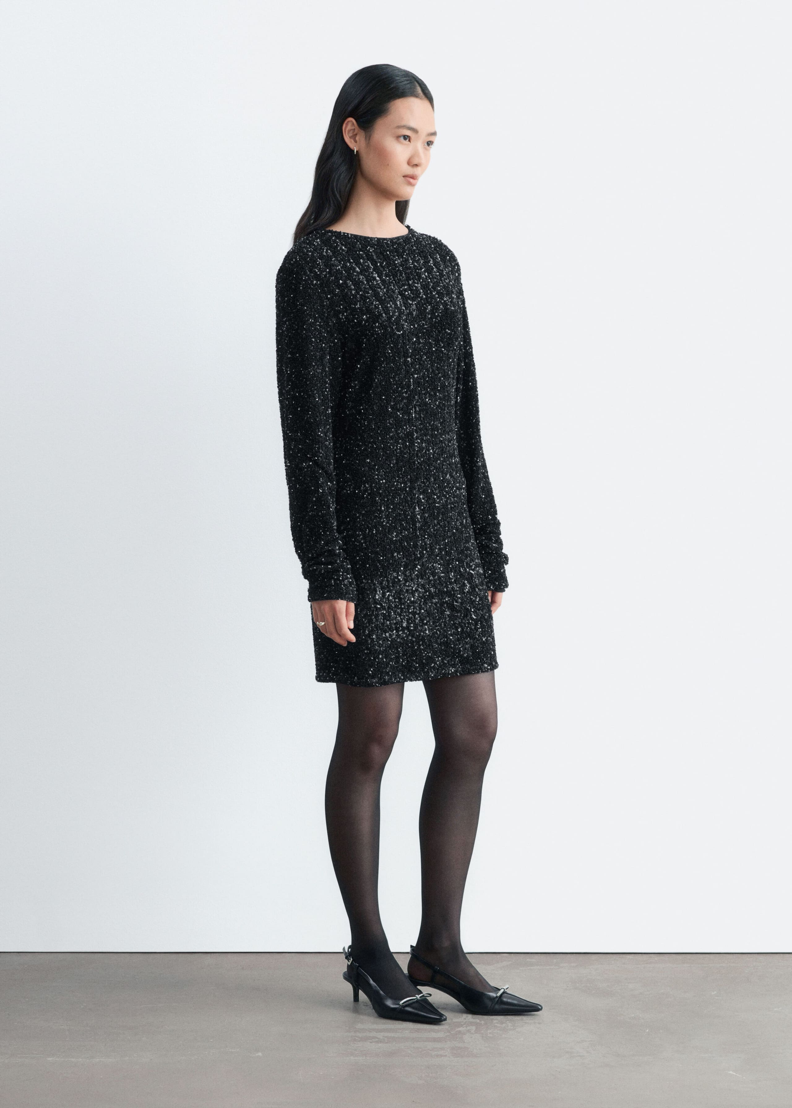 The model is wearing a short black sequined dress with long sleeves and a crew neckline. The look is styled with sheer black tights and black pointed-toe slingback heels.
