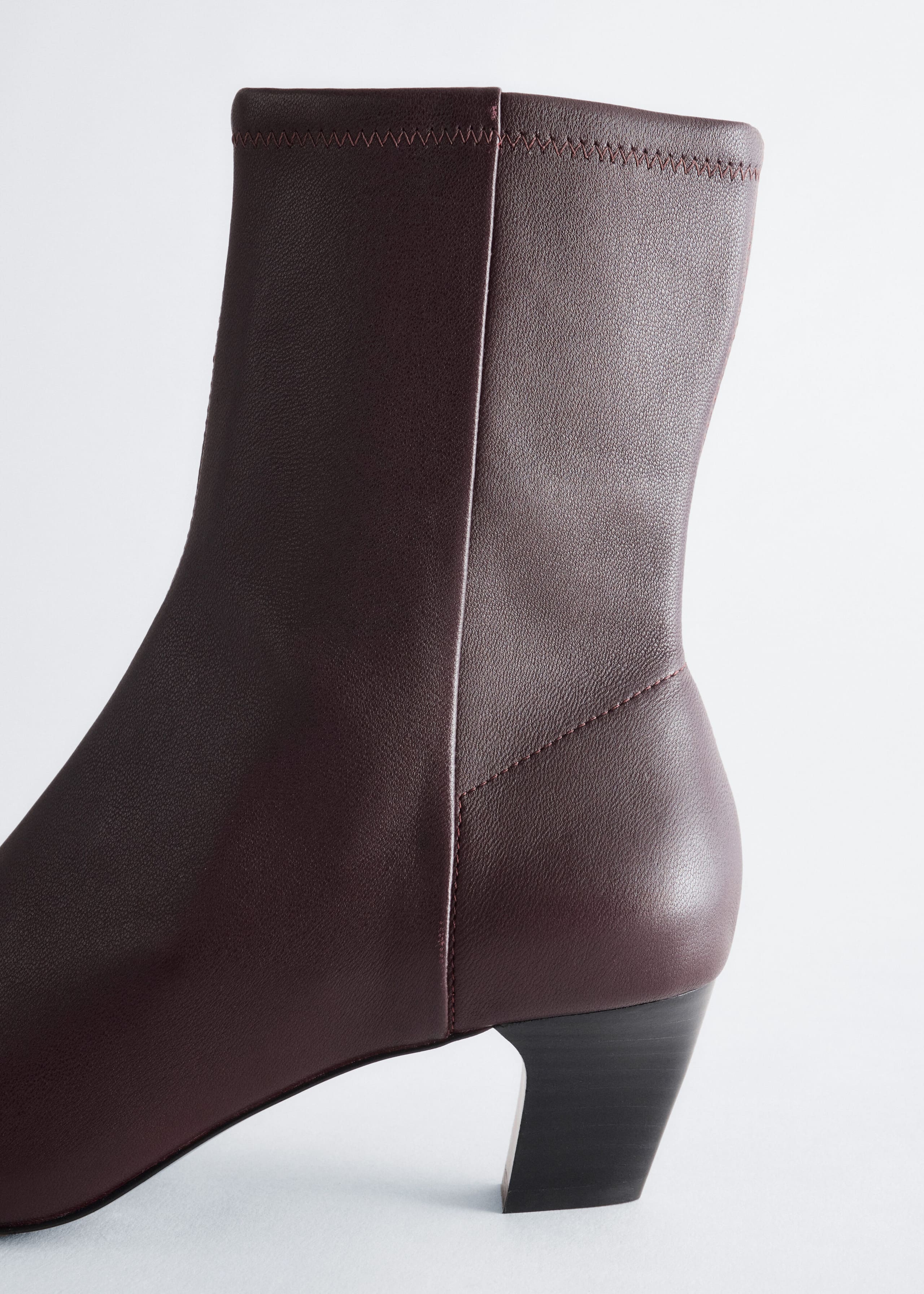Uno stivaletto alla caviglia in pelle color bordeaux scuro è mostrato in una vista laterale ravvicinata, con un tacco a blocco nero dalla forma curva e una cucitura verticale lungo il lato. Lo stivaletto presenta una texture liscia in pelle e un dettaglio con cucitura a zigzag lungo l’orlo superiore.