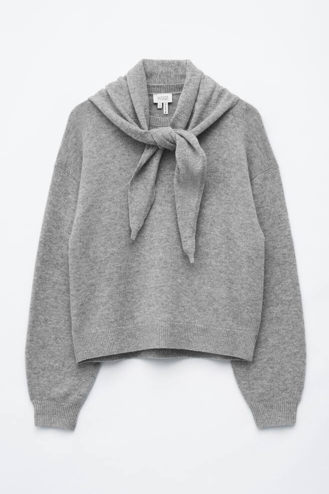 Ein hellgrauer Pullover aus Merinowolle mit Schal-Kragen liegt flach in frontaler Ansicht und prÃ¤sentiert einen integrierten Strickschal, der locker mittig auf der Brust geknotet ist. Der Pullover hat lange Ãrmel mit Ã¼berschnittenen Schultern, RippbÃ¼ndchen und einen Rippbund am Saum. - 1