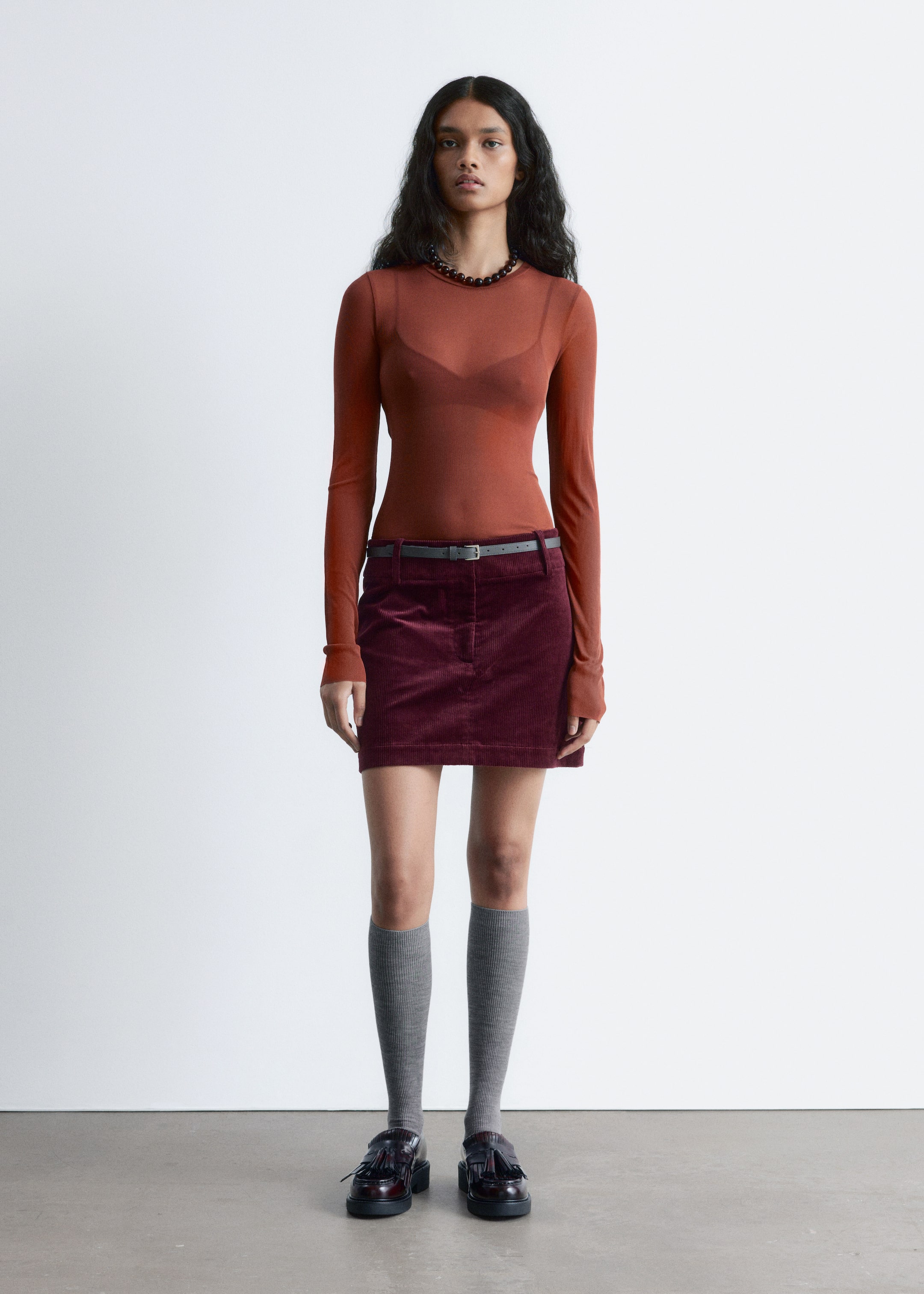 View larger image: A rust-colored, sheer long-sleeved top is being worn, revealing a darker bra underneath. A maroon corduroy mini skirt, featuring visible vertical ribs and belt loops, is also being worn with a thin dark belt.