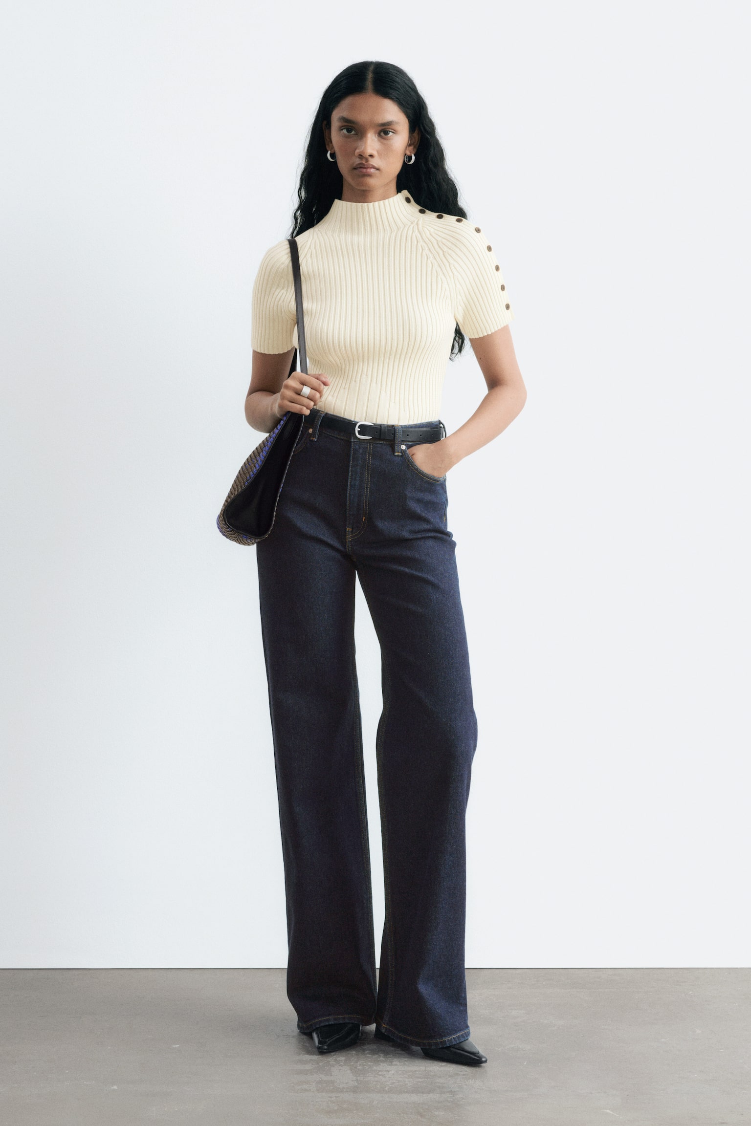 Die Model trÃ¤gt hoch geschnittene Wide-Leg-Jeans aus dunklem Denim, kombiniert mit einem schwarzen GÃ¼rtel und einem in die Hose gesteckten cremefarbenen Ripp-Top. Sie steht mit einer Hand in der Tasche und prÃ¤sentiert die lange, gerade Silhouette der Jeans. - 1