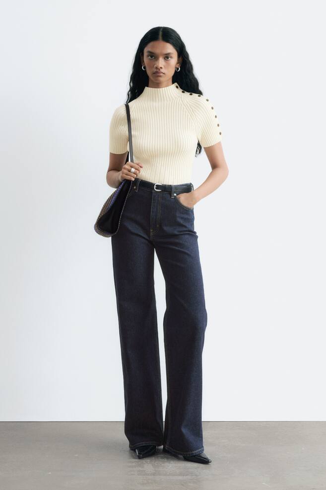 High-waisted, wide-leg jeans in dark wash denim are worn by the model, styled with a black belt and a tucked-in cream ribbed top. The model is standing with one hand in the pocket, showcasing the long, straight silhouette of the jeans. - 1