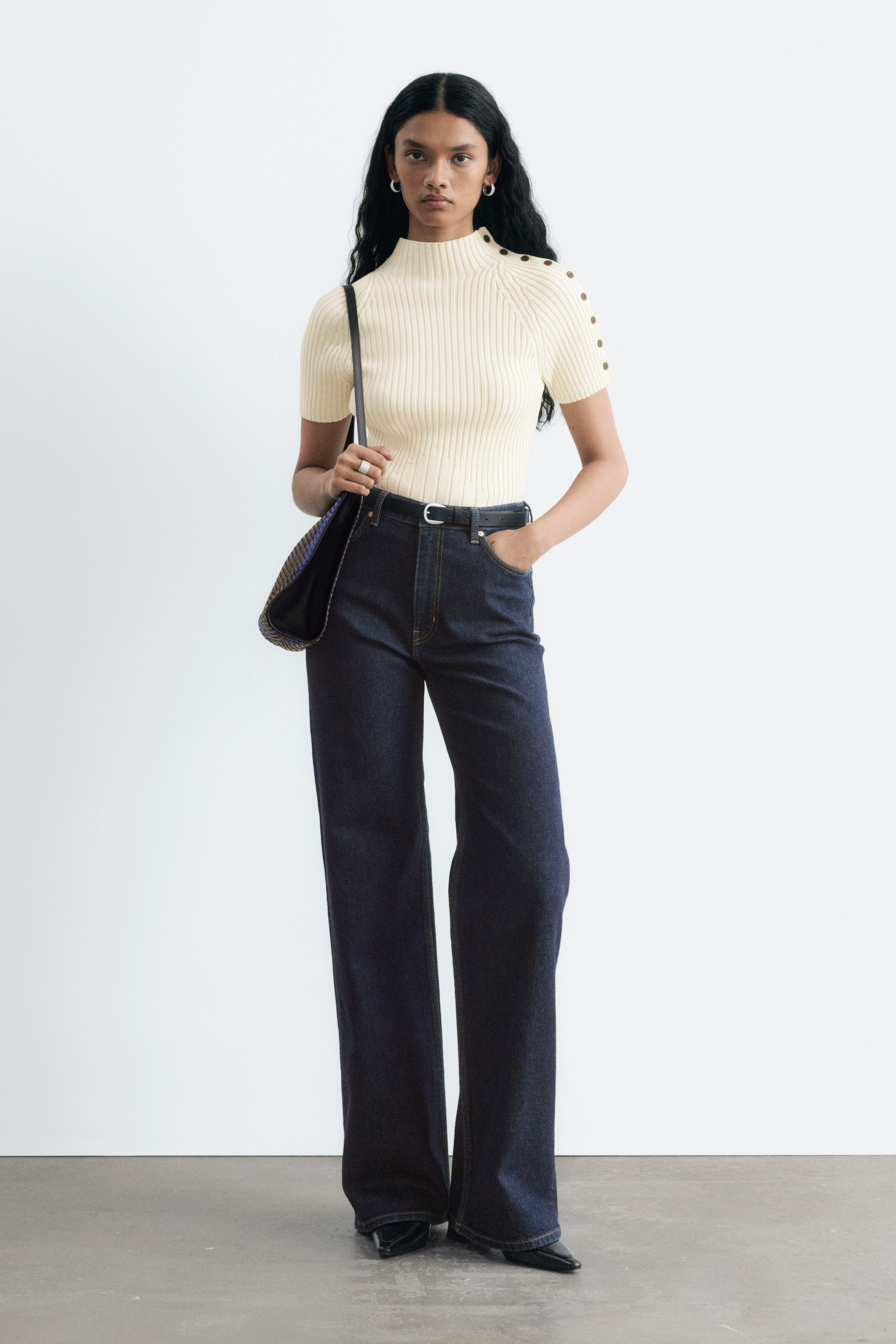 Visualizza immagine più grande: Jeans a vita alta, a gamba larga in denim scuro, indossati dalla modella, abbinati a una cintura nera e a un top a coste color crema infilato nei pantaloni. La modella è in piedi con una mano in tasca, mettendo in risalto la silhouette lunga e dritta dei jeans.