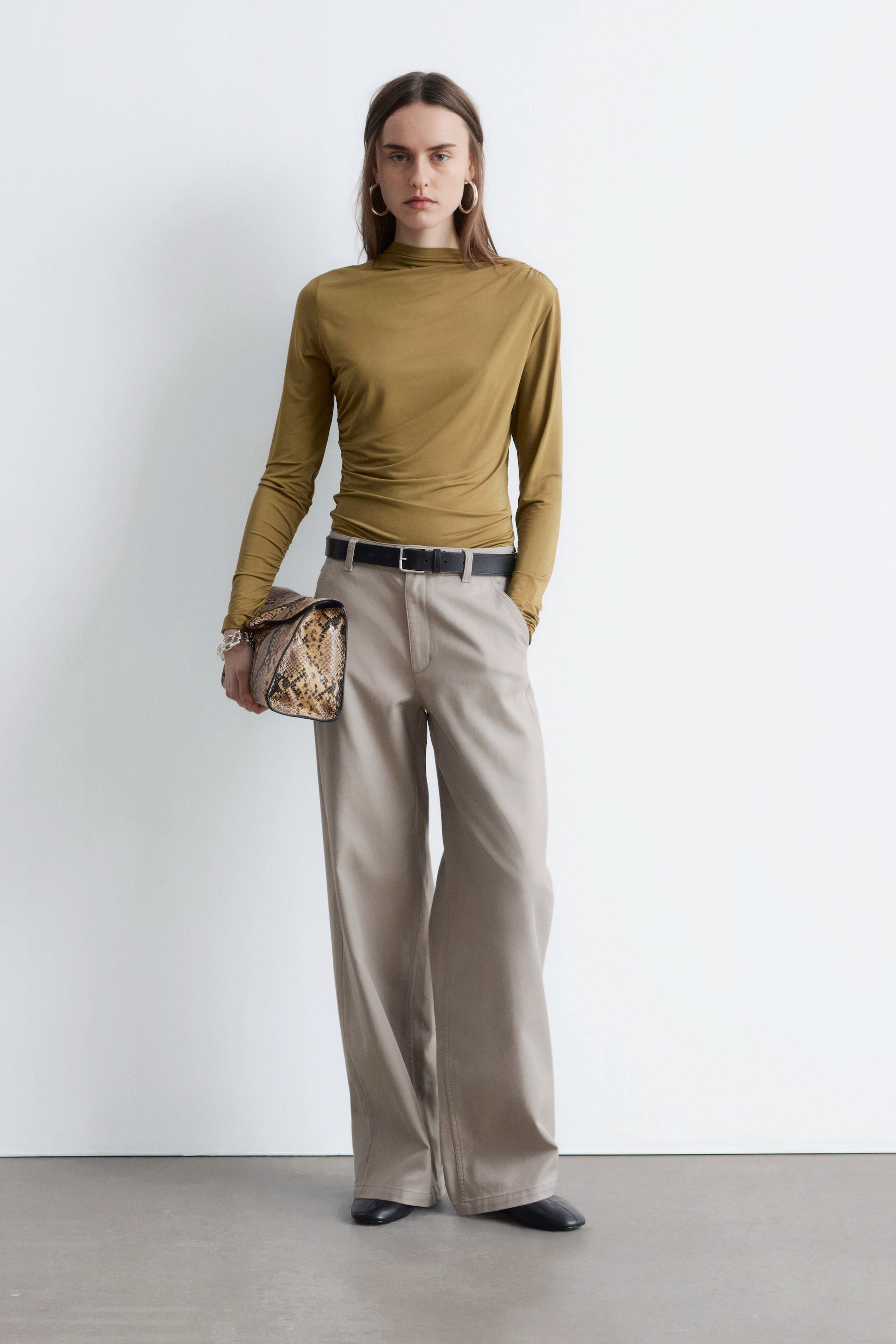 Se større billede: En olivengrøn mock-neck langærmet top med side-ruching bæres sammen med lyse beige bredbenede bukser. Bukserne er strammet i taljen med et sort bælte, og begge beklædningsgenstande vender fremad og præsenterer en afslappet pasform.