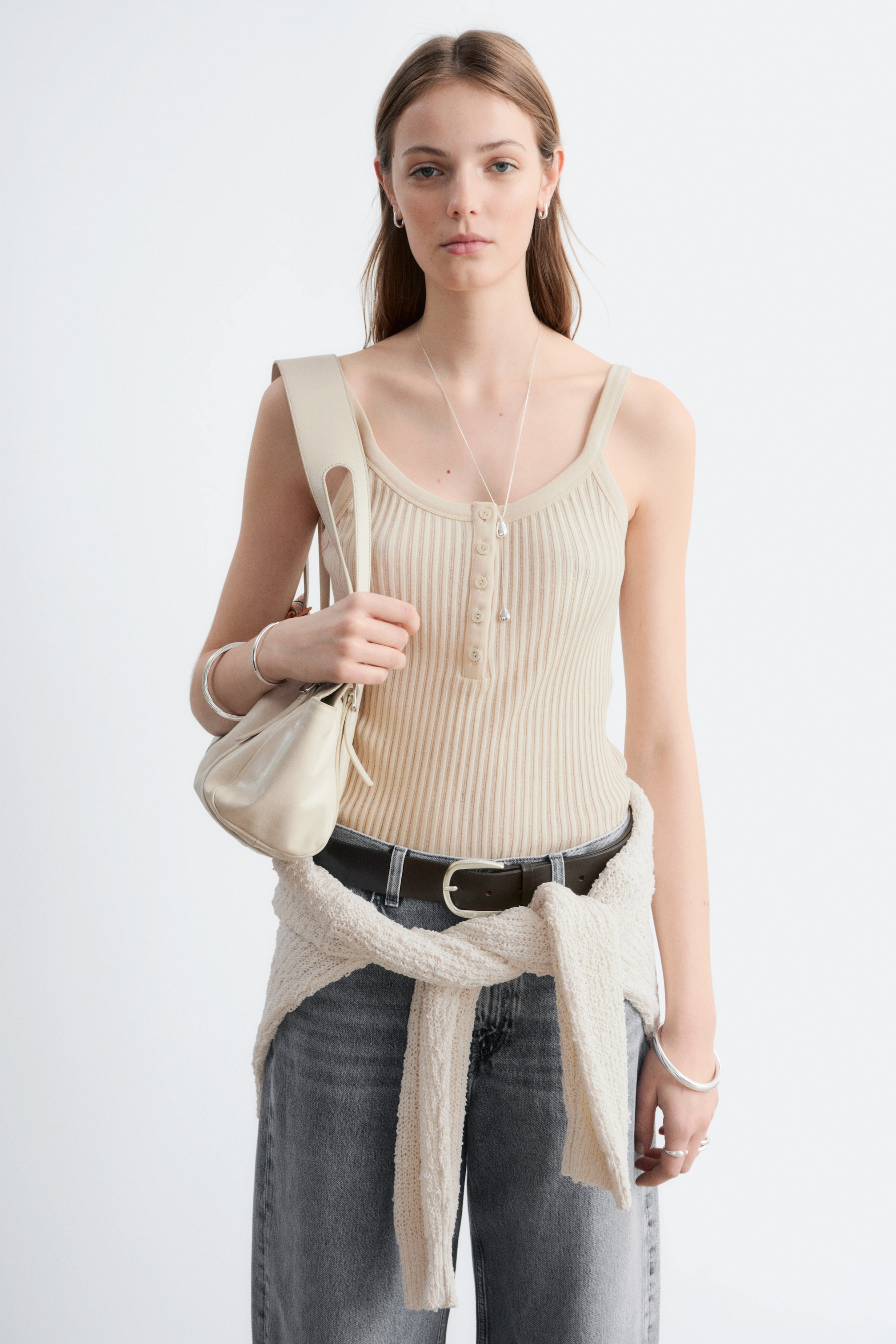 View larger image: The model is wearing a light-coloured ribbed vest top, featuring a scoop neckline, thin shoulder straps, and a half-button placket. The top is tucked into dark jeans, with a light jumper tied around the model's waist.