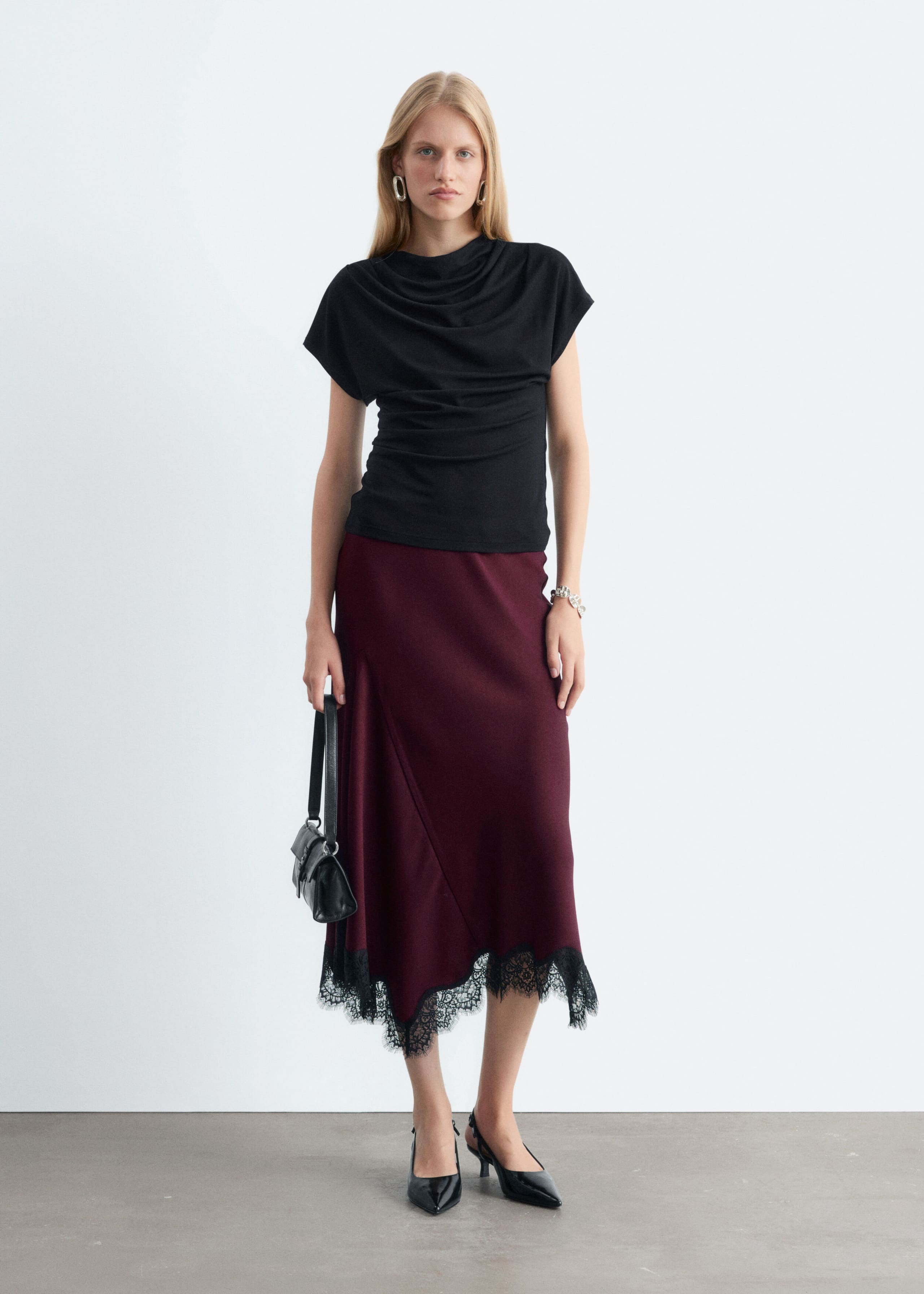 A burgundy midi skirt with a flared silhouette and an asymmetric hemline trimmed in black lace is worn by the model. The skirt is styled with a draped black top and black slingback heels, while a small black handbag is carried.