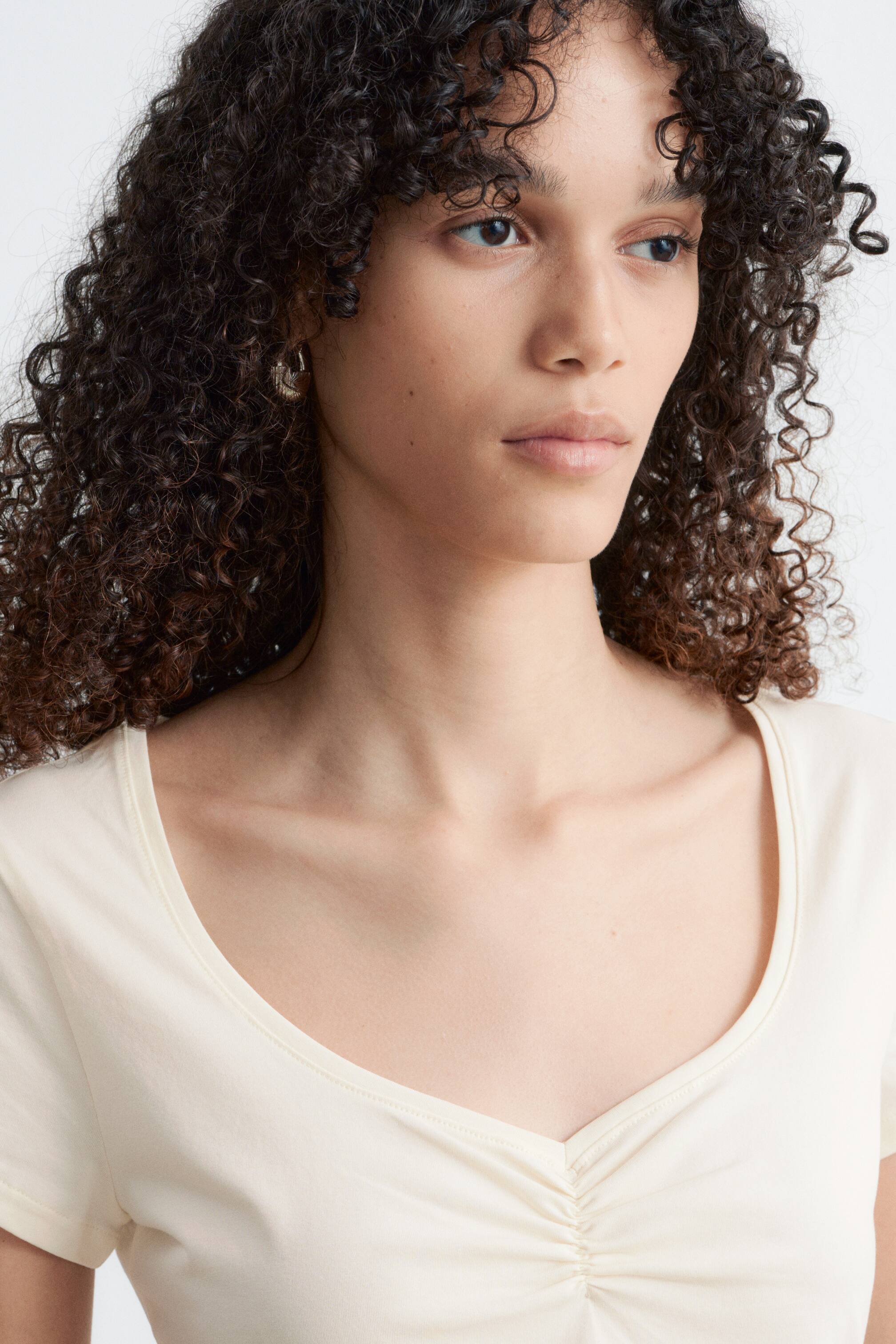 Grotere afbeelding bekijken: Een crèmekleurige top met korte mouwen wordt vanuit een voor-rechterhoek getoond, met een gescoopt V-halslijn en plooien aan de voorkant.