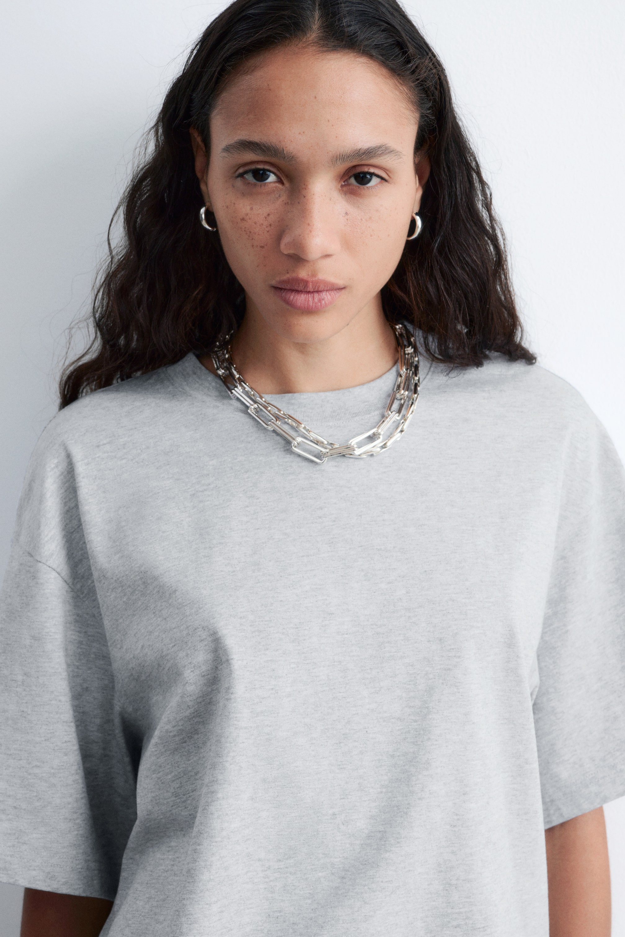 Grotere afbeelding bekijken: Een lichtgrijs T-shirt met ronde hals wordt van voren gepresenteerd, met korte mouwen en een ontspannen pasvorm. Een gelaagde zilverkleurige ketting, gemaakt van rechthoekige paperclip schakels, rust op de halslijn van het T-shirt.