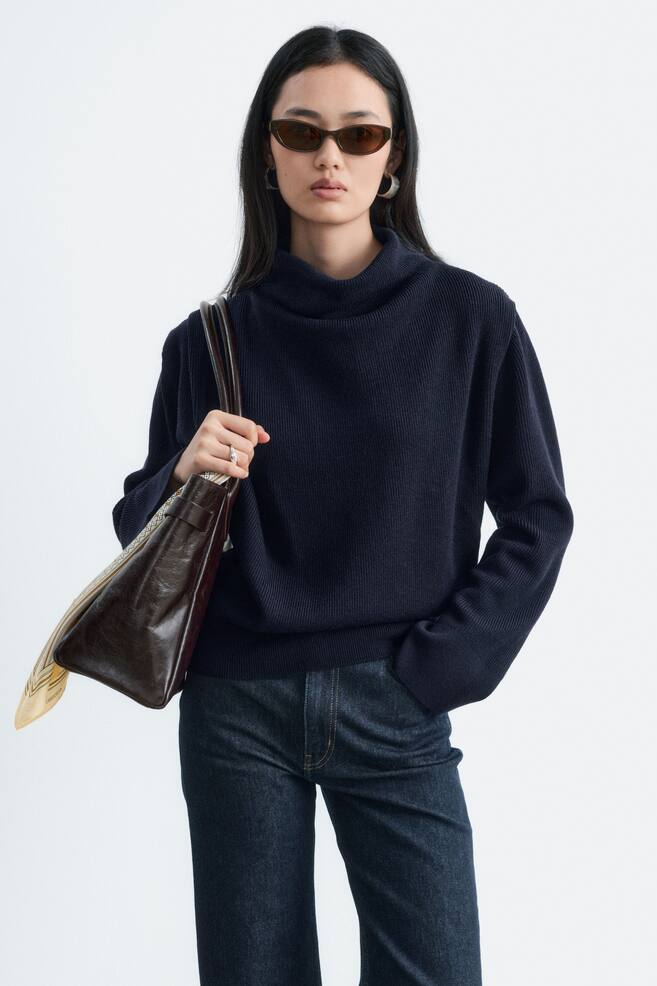 A navy blue ribbed wool jumper featuring a high, draped cowl neckline and long sleeves is worn by the model. The jumper is styled with dark denim jeans, with the model carrying a dark brown leather shoulder bag over one arm. - 1