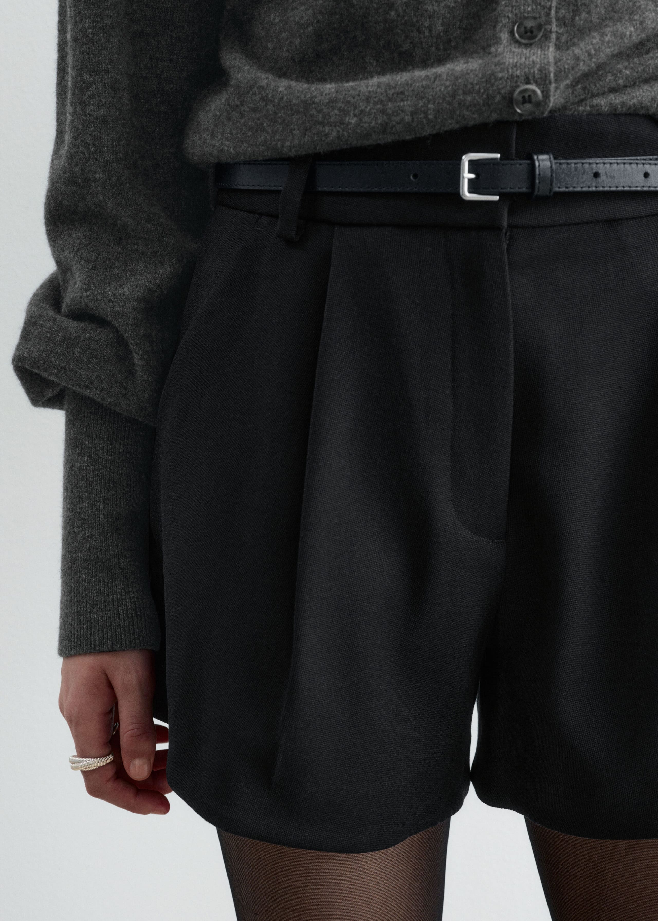 Black high-waisted shorts featuring front pleats and a slim black leather belt with a silver square buckle are being worn by the model. The shorts are styled with a tucked-in grey knit cardigan.