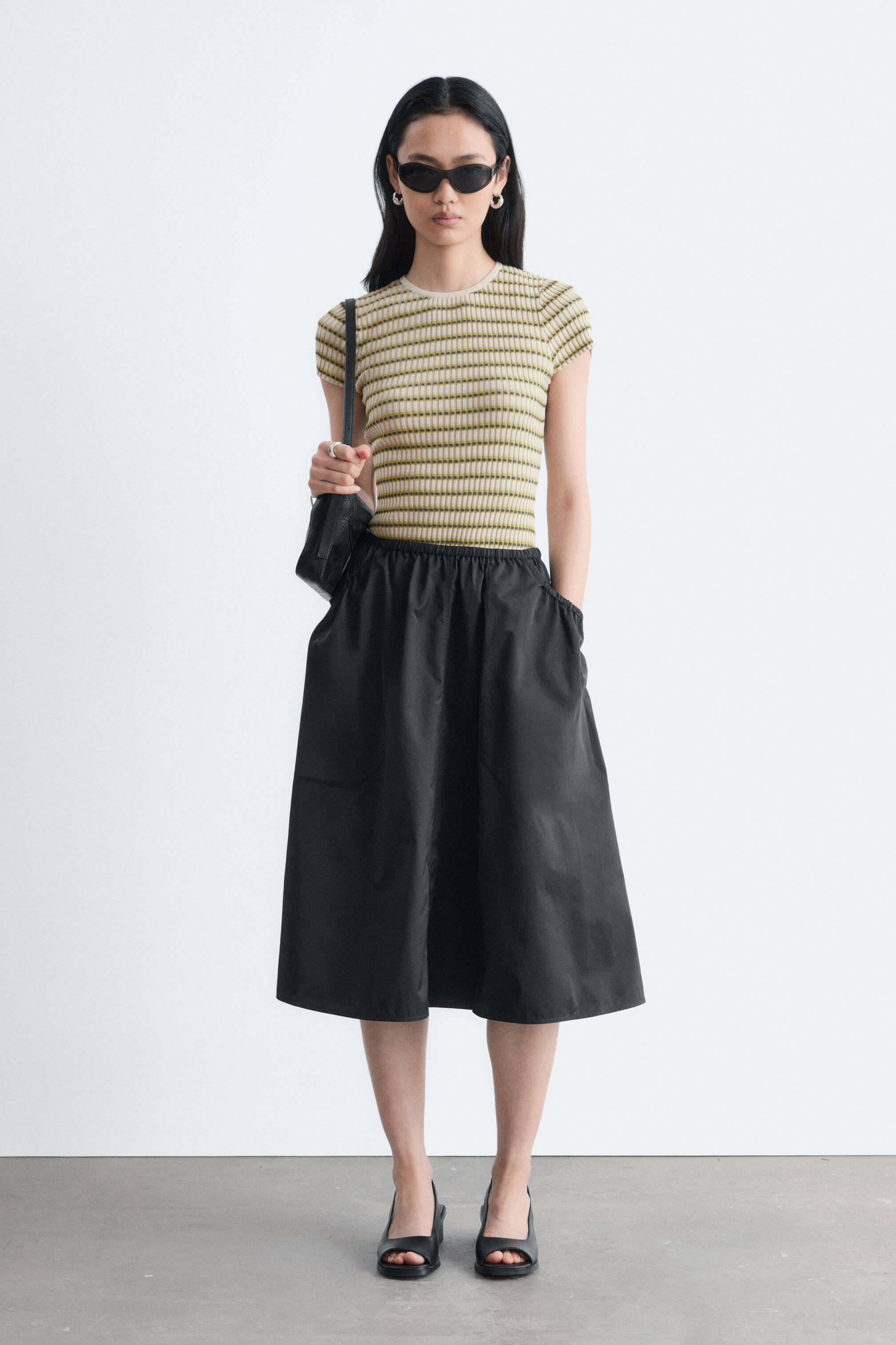 Zobrazit větší obrázek: Černá midi sukně se vyznačuje elastickým, nařaseným pasem a rozšiřuje se do áčkové siluety. Obsahuje viditelné boční kapsy a sahá do poloviny lýtek.