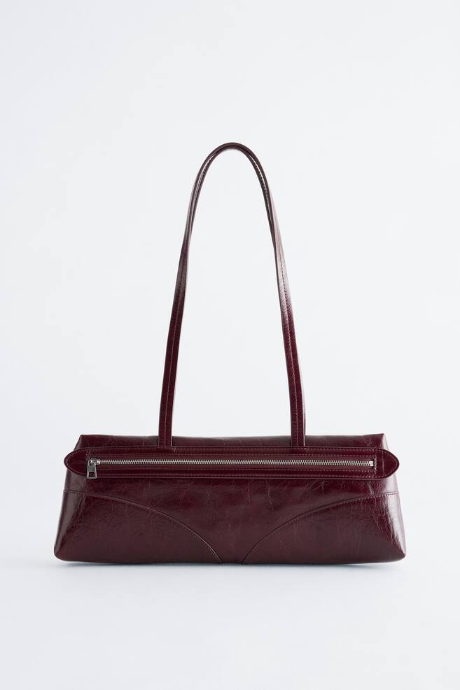 Un sac bandouliÃ¨re en cuir bordeaux foncÃ© est prÃ©sentÃ© debout de face, mettant en valeur une silhouette rectangulaire allongÃ©e et discrÃ¨te, Ã  la finition brillante et plissÃ©e. Le sac affiche de longues bandouliÃ¨res fines ainsi quâune fermeture zippÃ©e horizontale ton argent le long dâun panneau frontal surÃ©levÃ©, avec des surpiqÃ»res courbes prÃ¨s de la base. - 1