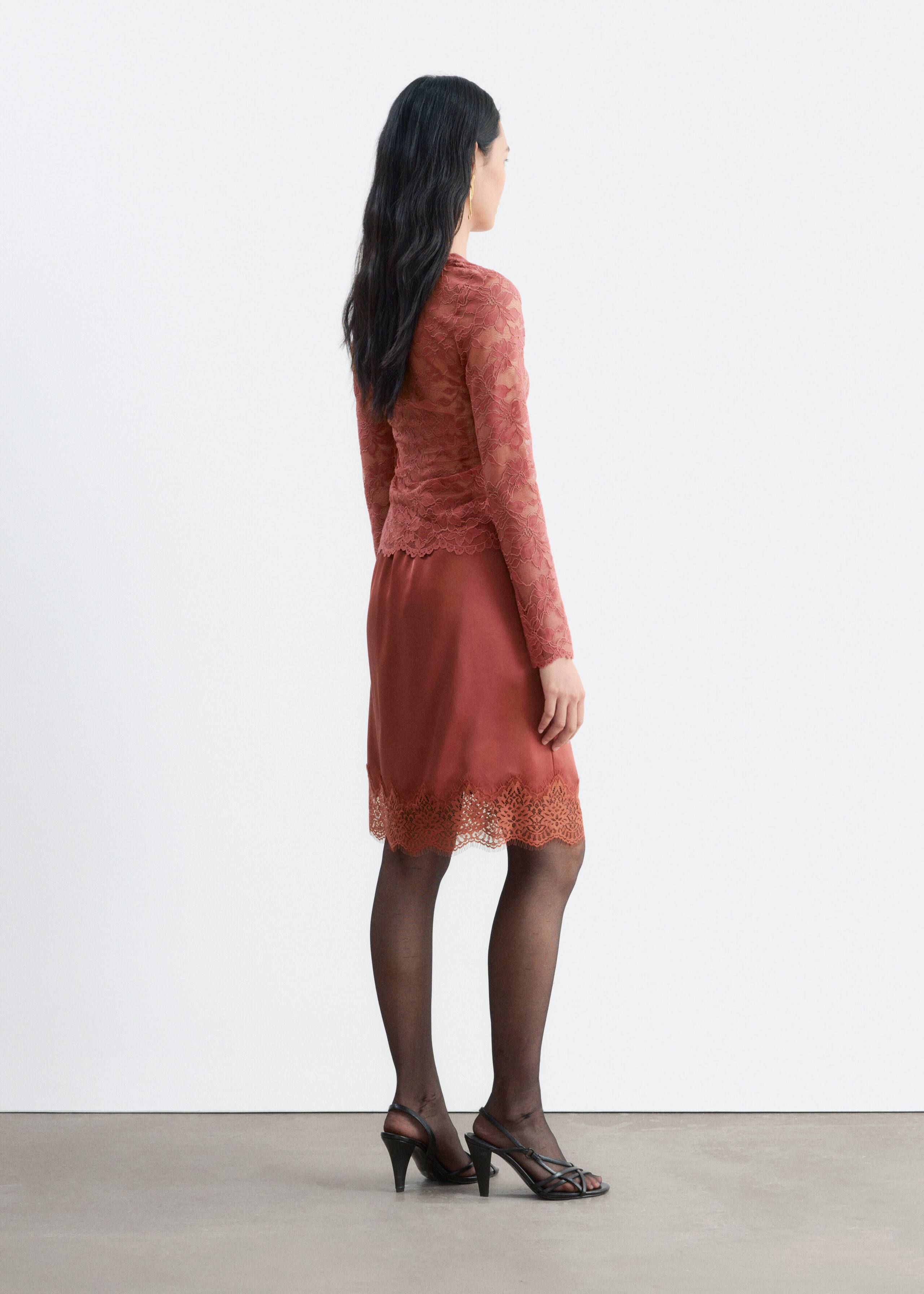 The model is wearing a rust-coloured, long-sleeved lace top and a matching knee-length satin skirt with a wide lace-trimmed hem. The ensemble is styled with sheer black tights and black strappy heeled sandals.