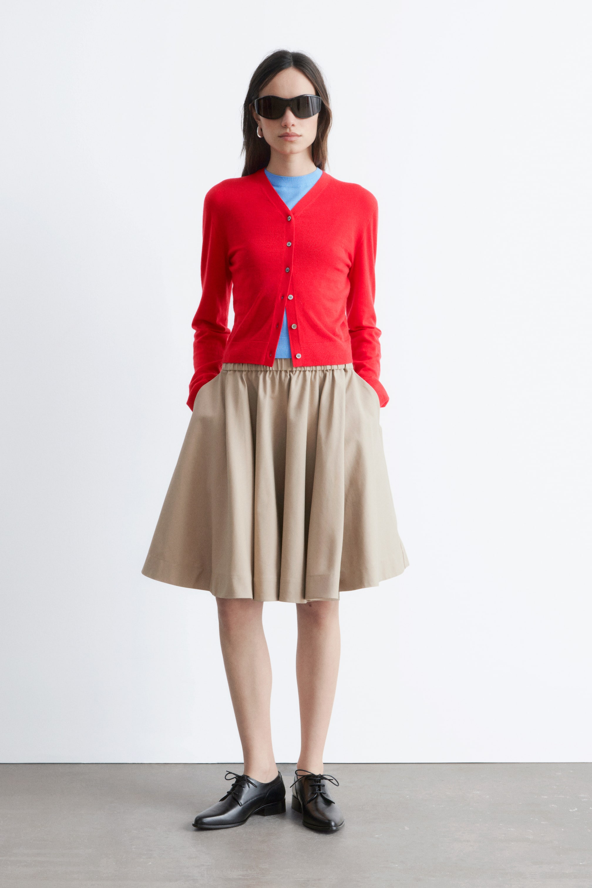 Näytä suurempi kuva: Punainen V-pääntiellä varustettu neuletakki, jossa on nappikiinnitys ja pitkät hihat, on kerrostettu sinisen V-pääntiellä olevan yläosan päälle. Se on yhdistetty polvipituiseen, beigeen A-linjaiseen hameeseen, jossa on joustava kerätty vyötärö ja sivutaskut, sekä mustiin nauhallisiin kenkiin.