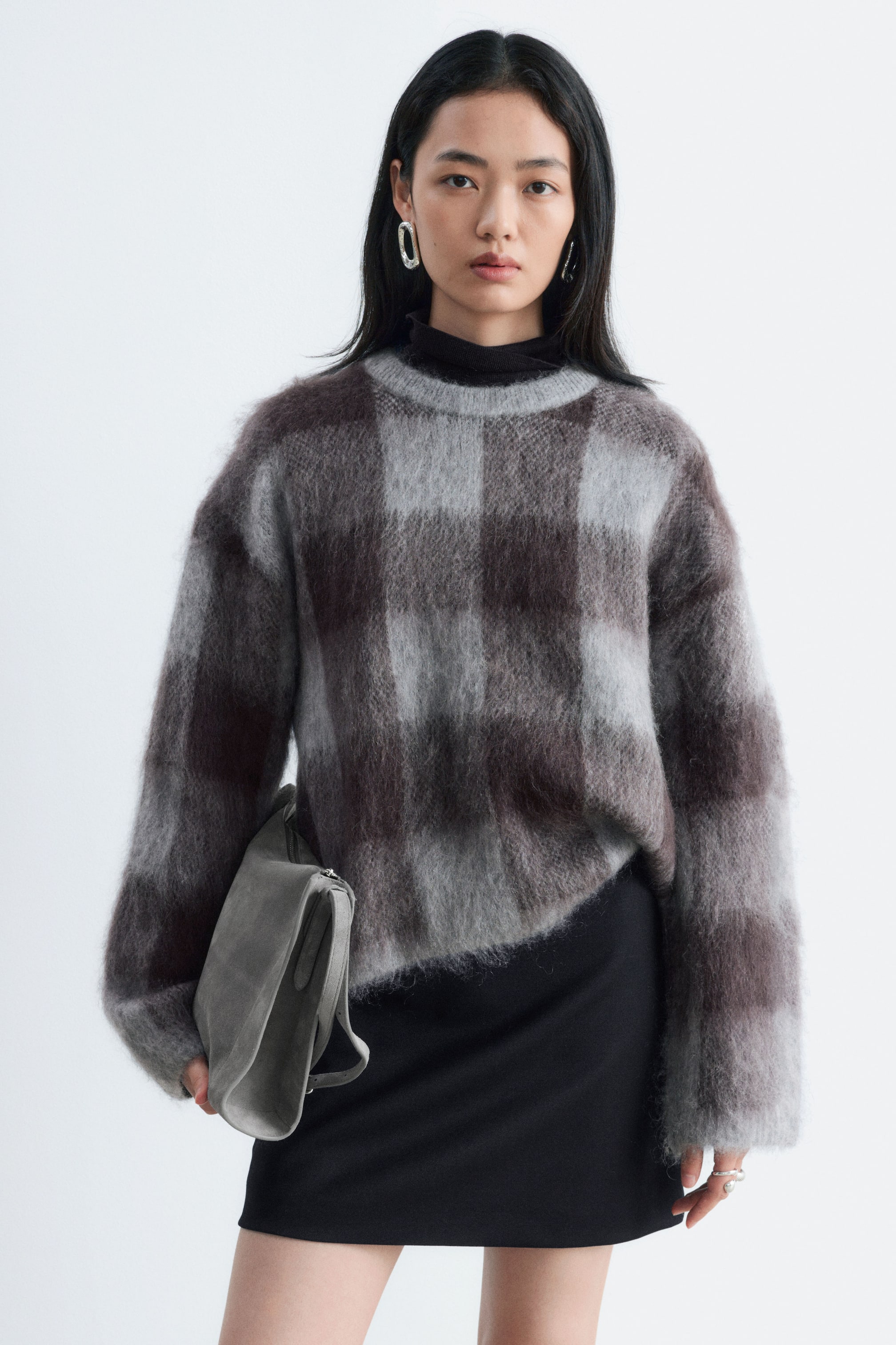 Ver imagen más grande: La modelo lleva un jersey de mezcla de mohair con textura afelpada y un estampado de cuadros jacquard cepillado en gris y marrón, sobre un cuello alto negro y una falda negra. La modelo sostiene bajo un brazo un bolso de ante gris.