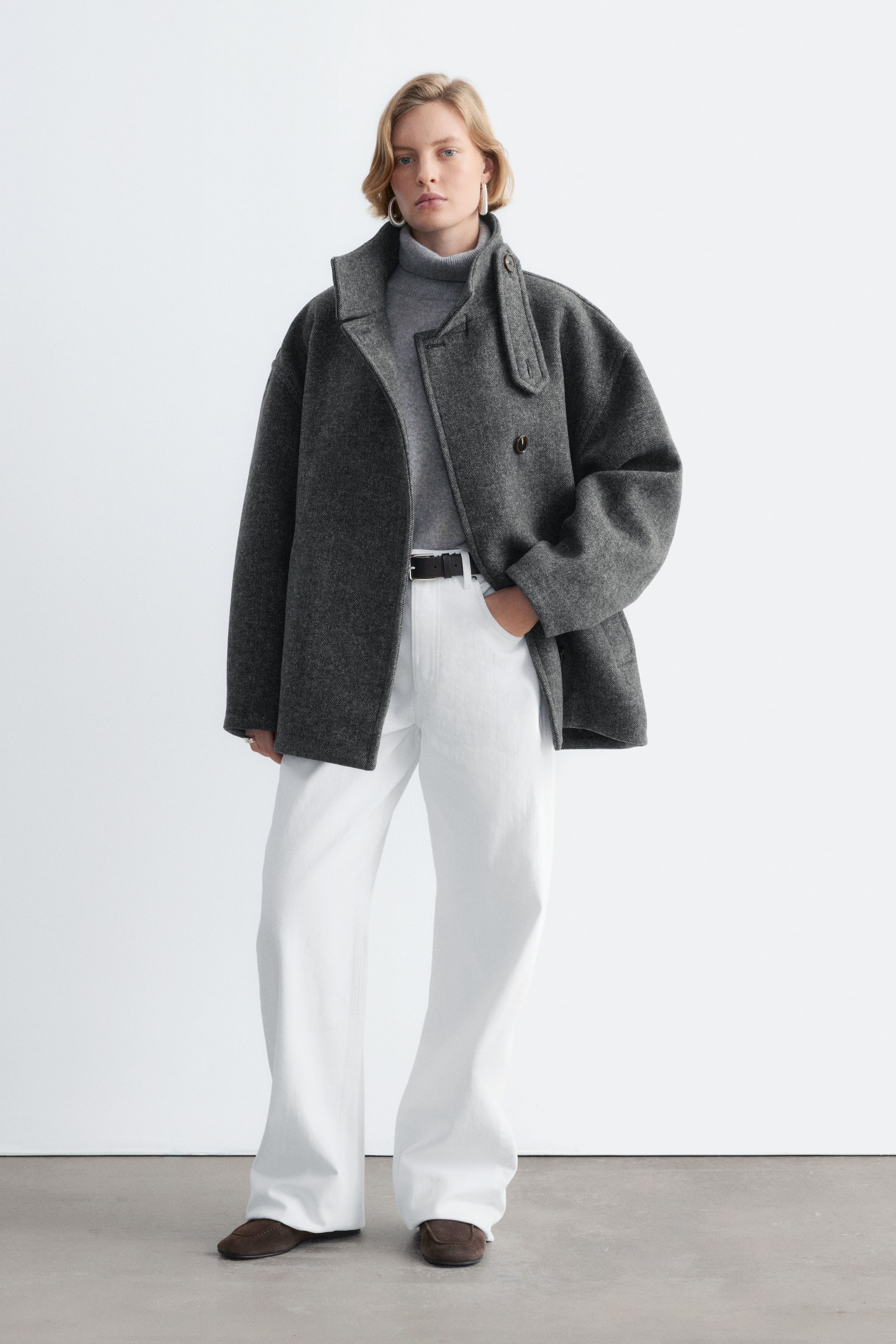 Visualizza l'immagine ingrandita: Un cappotto grigio scuro in misto lana con motivo pied-de-poule, caratterizzato da una silhouette oversize, maniche raglan e un colletto alto con linguetta, è indossato aperto dalla modella. Il cappotto è abbinato come strato sopra un maglione dolcevita grigio e pantaloni bianchi con cintura nera.