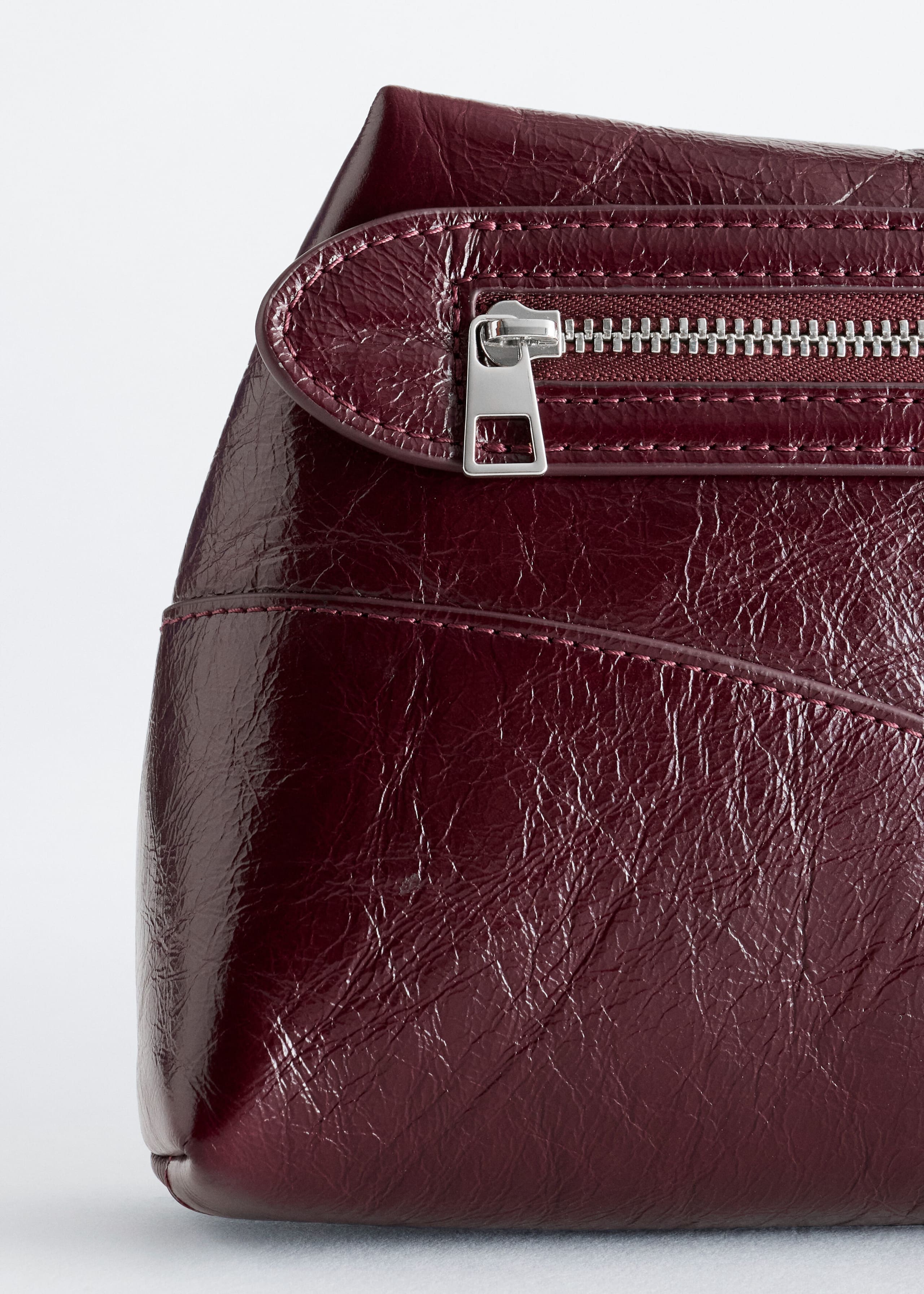 A close-up of a burgundy leather shoulder bag showcasing its glossy, crinkled texture and tonal stitching. A silver-tone metal zip with a rectangular pull tab is integrated into a stitched horizontal panel.