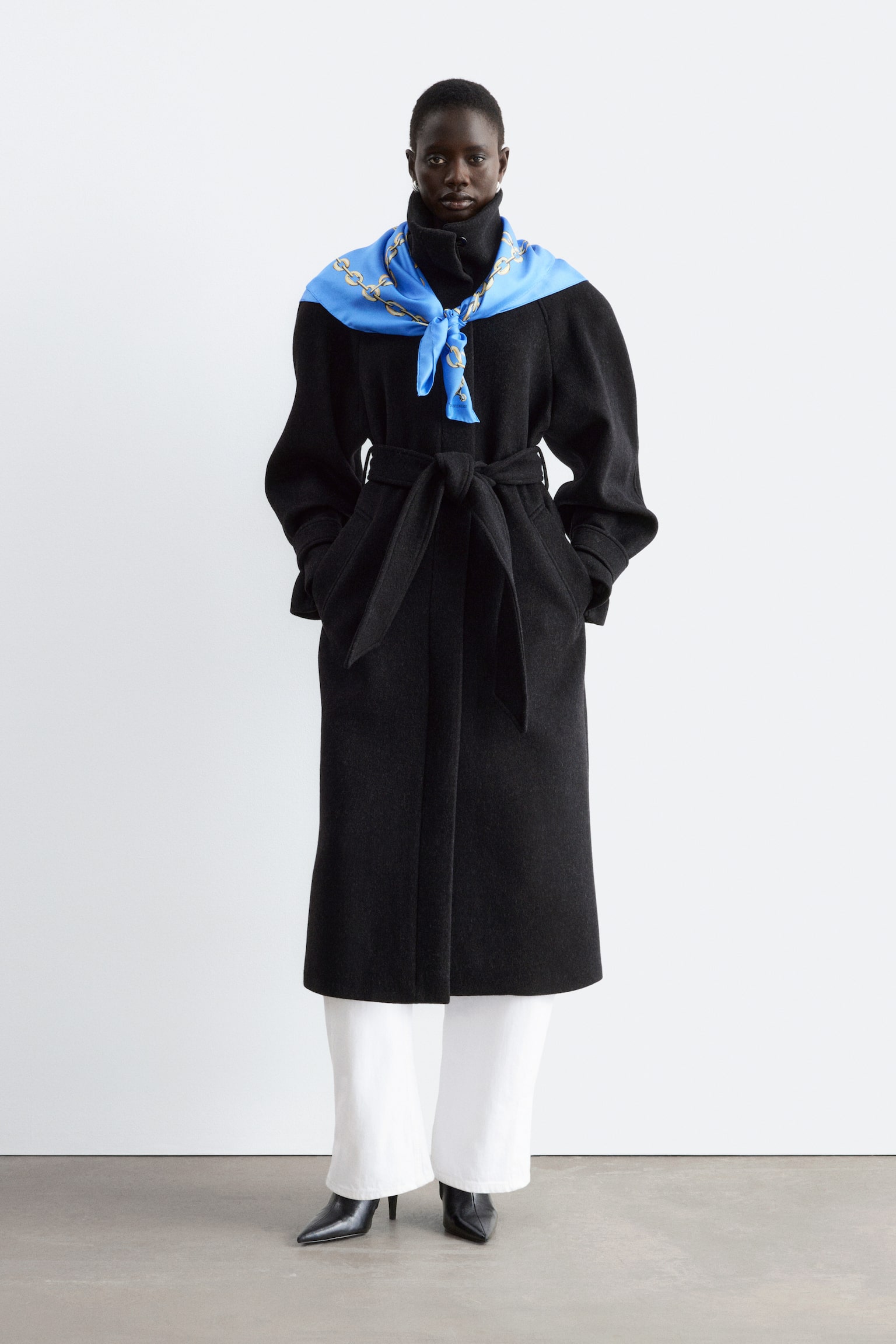 Un long manteau noir ceinturé, aux épaules structurées et à col montant, est porté par le mannequin. Ce dernier pose les mains dans les poches du manteau, en accessoirisant la tenue avec une écharpe bleue à motifs nouée autour du col, un pantalon blanc et des bottes noires. - 1
